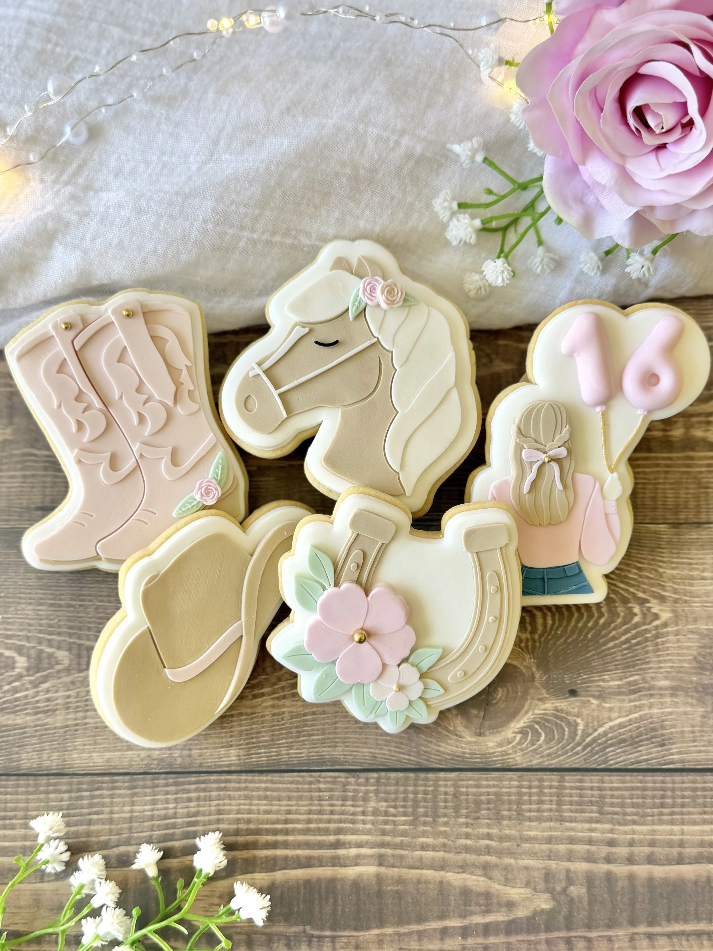 Western-themed 16th birthday cookies featuring a horse, cowgirl, pink cowboy boots, cowboy hat and floral horseshoe in soft pink, cream and brown