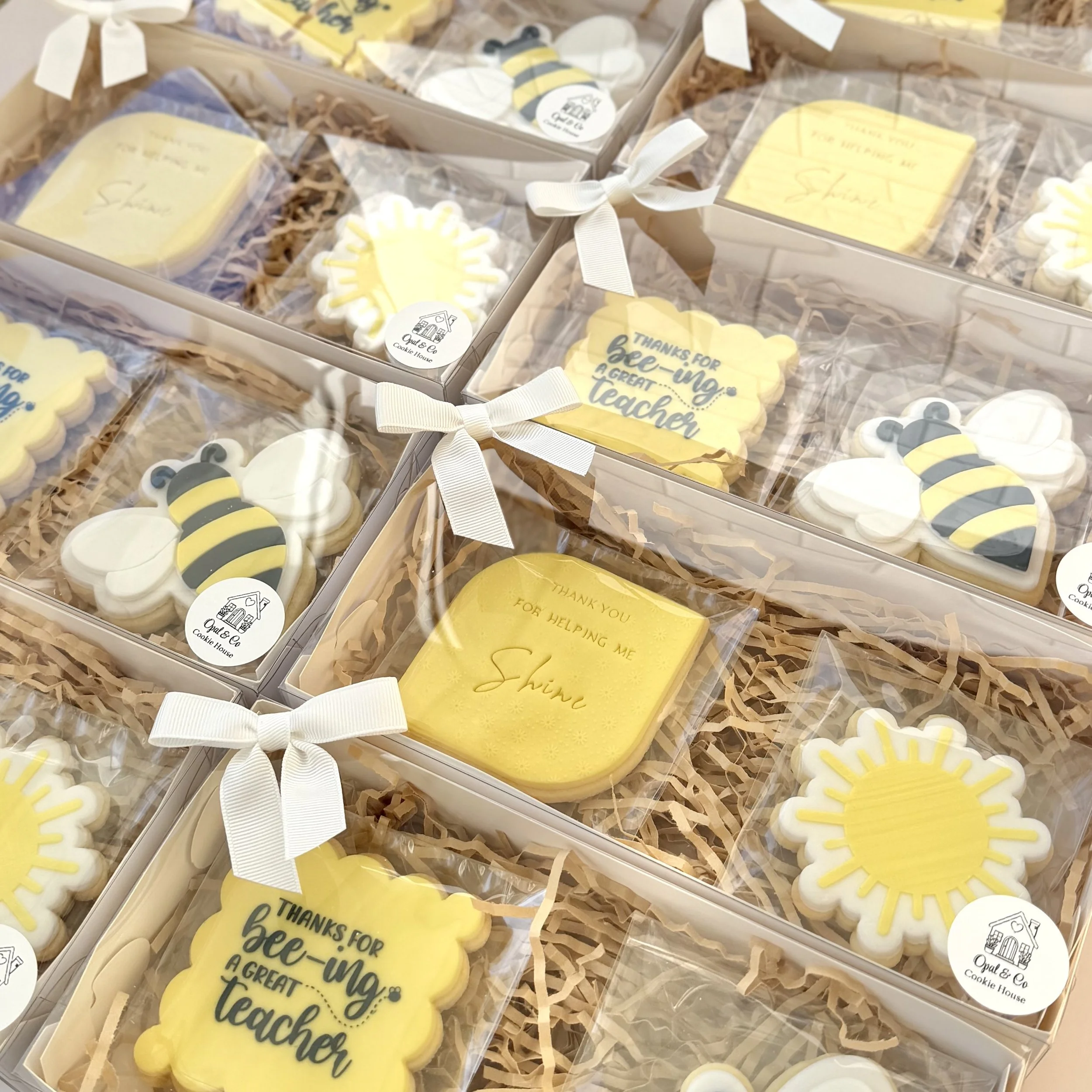 A collection of decorated cookies with bee and sunflower themes, packaged in clear boxes with white ribbons, placed on shredded paper in a display box