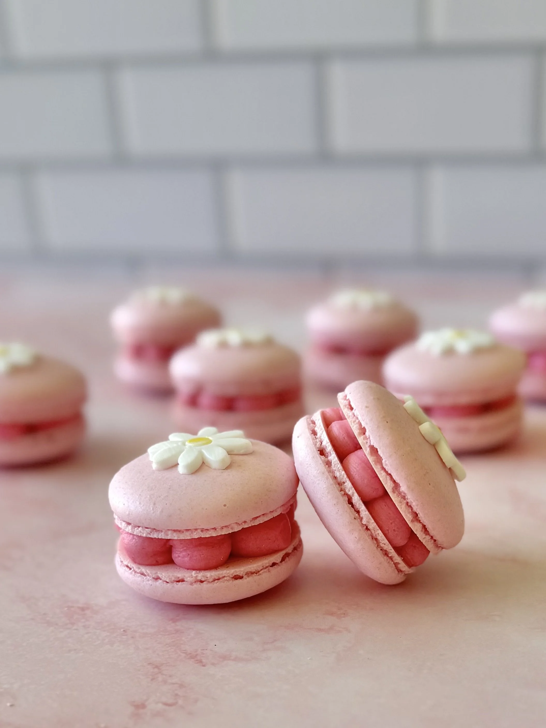 Close-up of pink macarons decorated with white flower-shaped icing on a light surface, with more macarons blurred in the background
