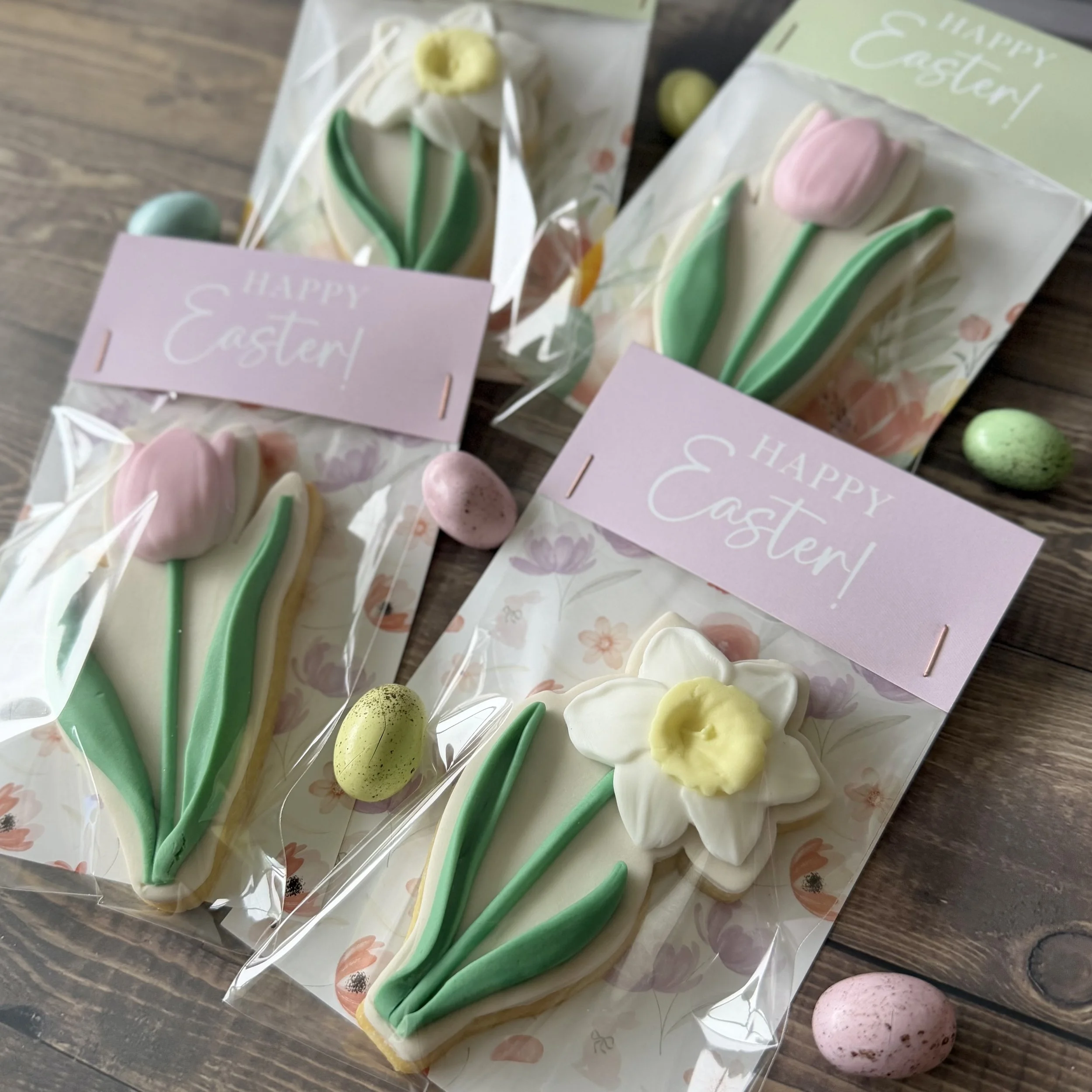 Decorative Easter cookies shaped like tulips and daffodils with pastel green stems and pink and yellow blossoms, packaged in clear plastic bags with pink and green tags that read 'Happy Easter!'
