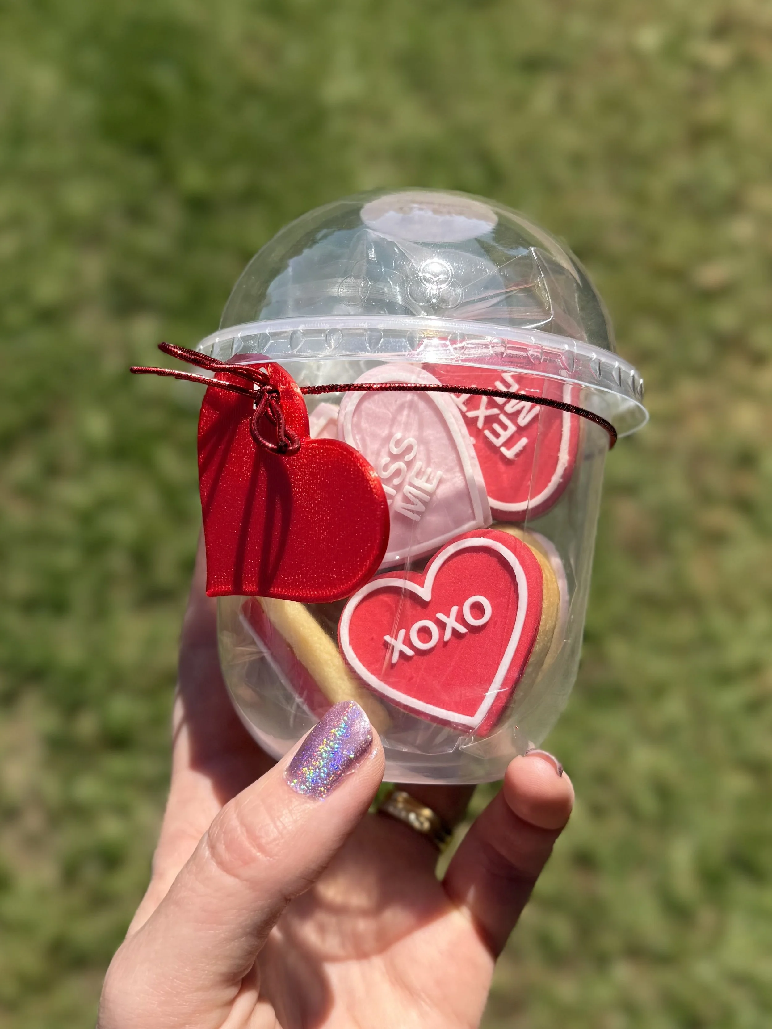 A hand holding a clear plastic container with Valentine's Day cookies inside, decorated with heart-shaped stickers and red ribbon