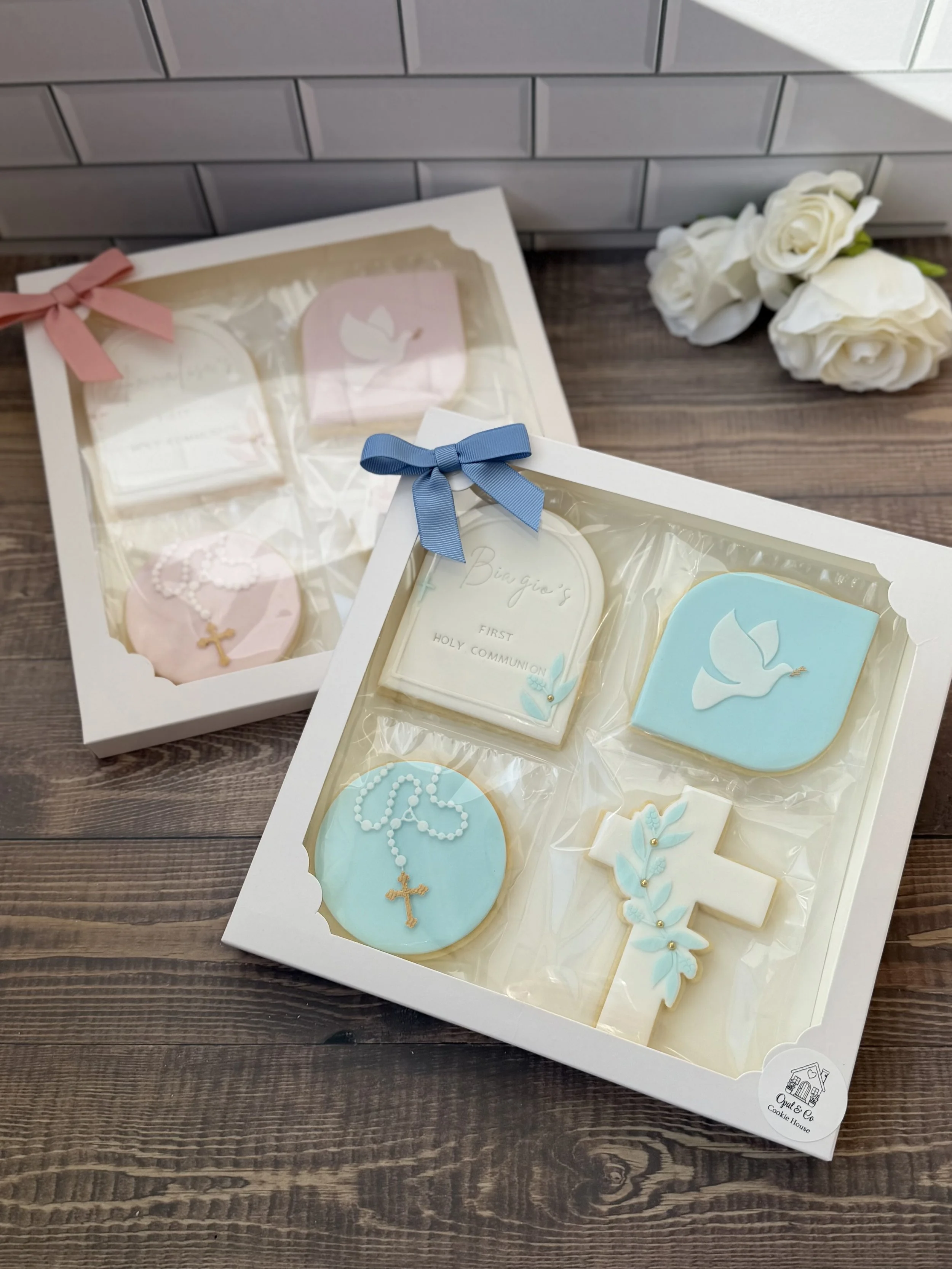 Two boxes of religious cookies decorated for a Holy Communion, one with pink and the other with blue fondant icing