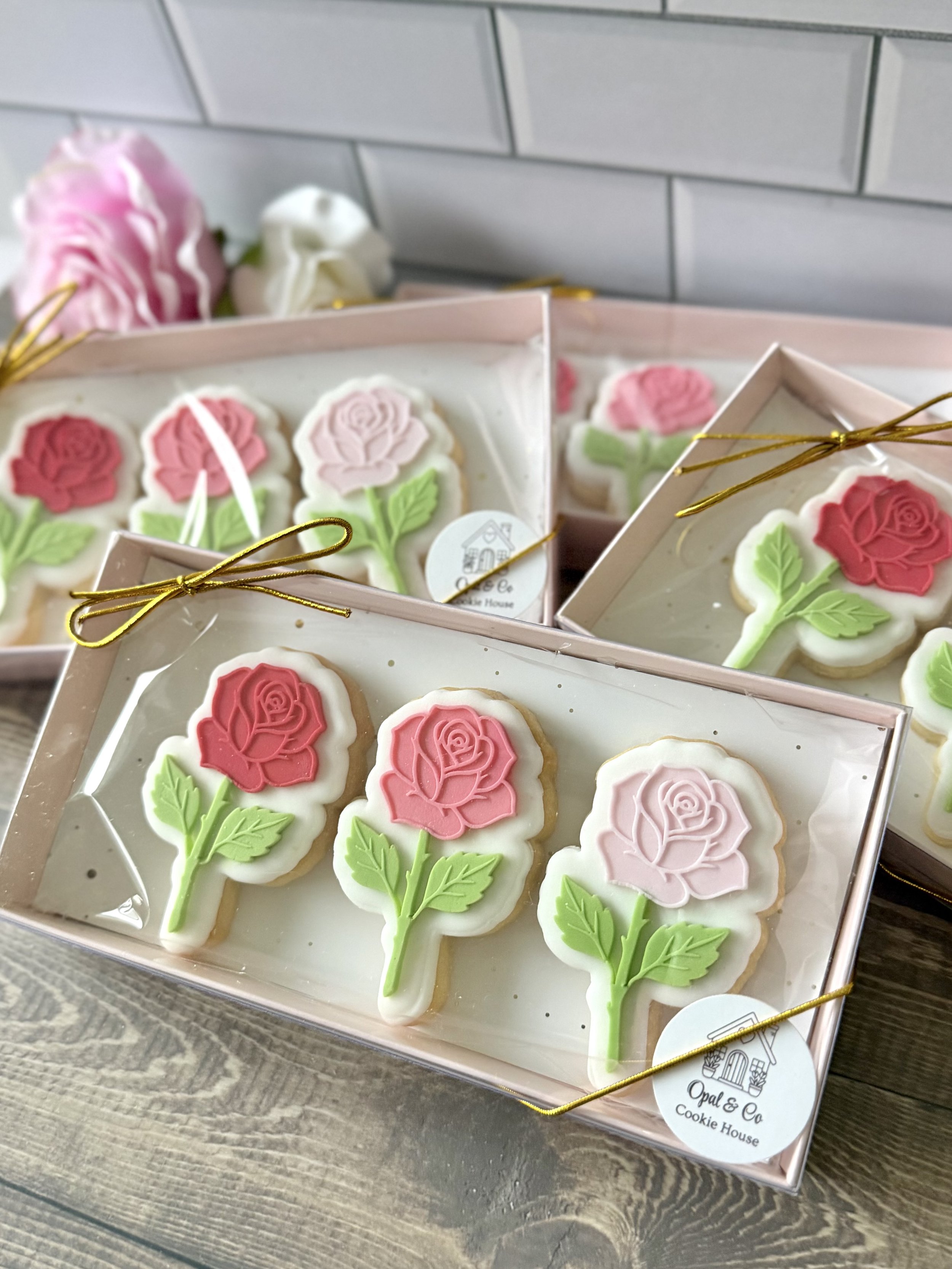 Decorative cookies shaped like pink and white roses with green leaves, arranged in pink boxes with gold ribbon, on a rustic wooden surface
