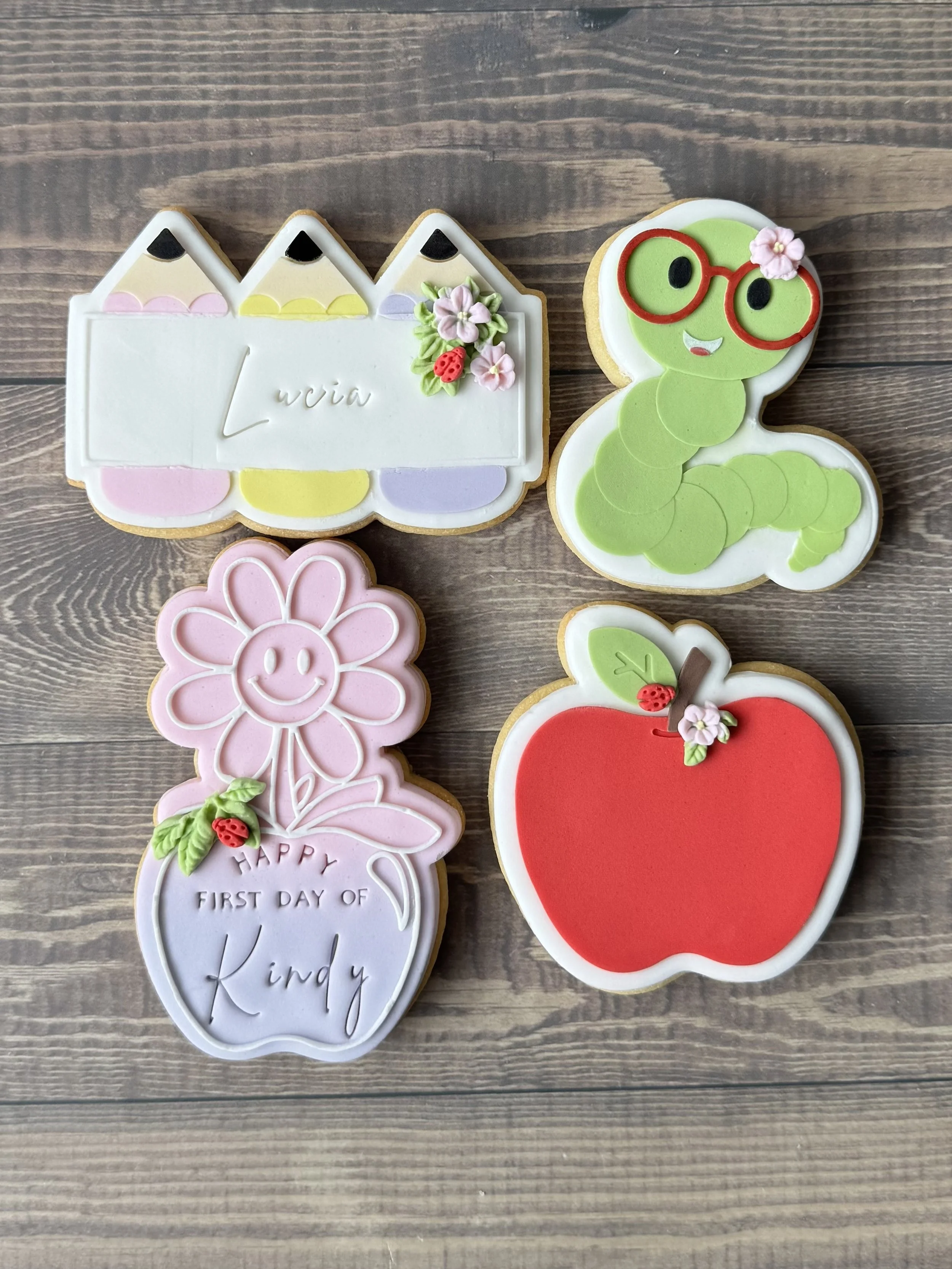 Decorated cookies including a pink, smiling flower with the text "HAPPY FIRST DAY OF KINDY", a caterpillar with green fondant and red glasses, and an apple with a pink flower and ladybug decoration
