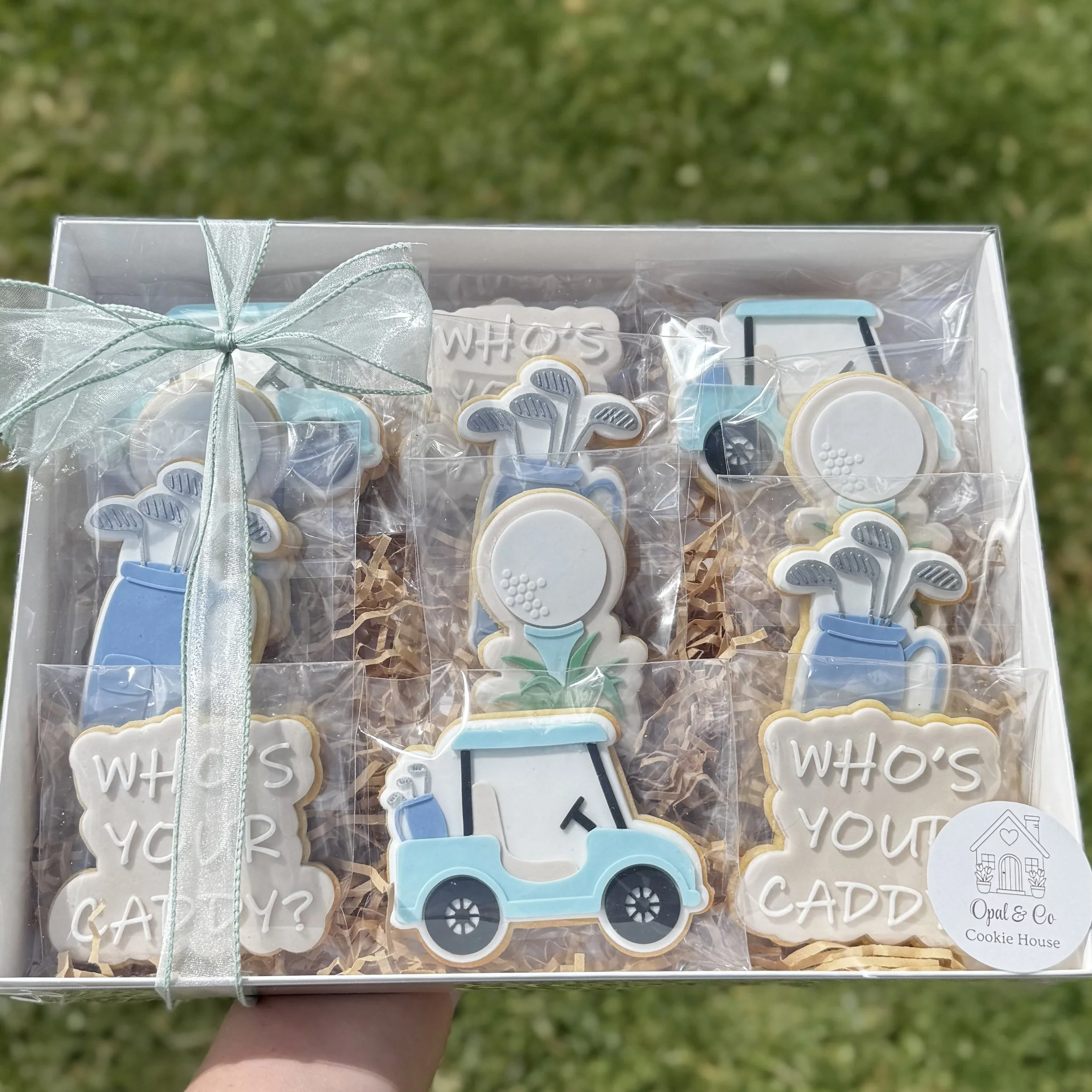 A gift box contains decorated cookies in a golf theme, with the words 'who's your caddy?', a golf cart, golf clubs and a golf tee with golf ball