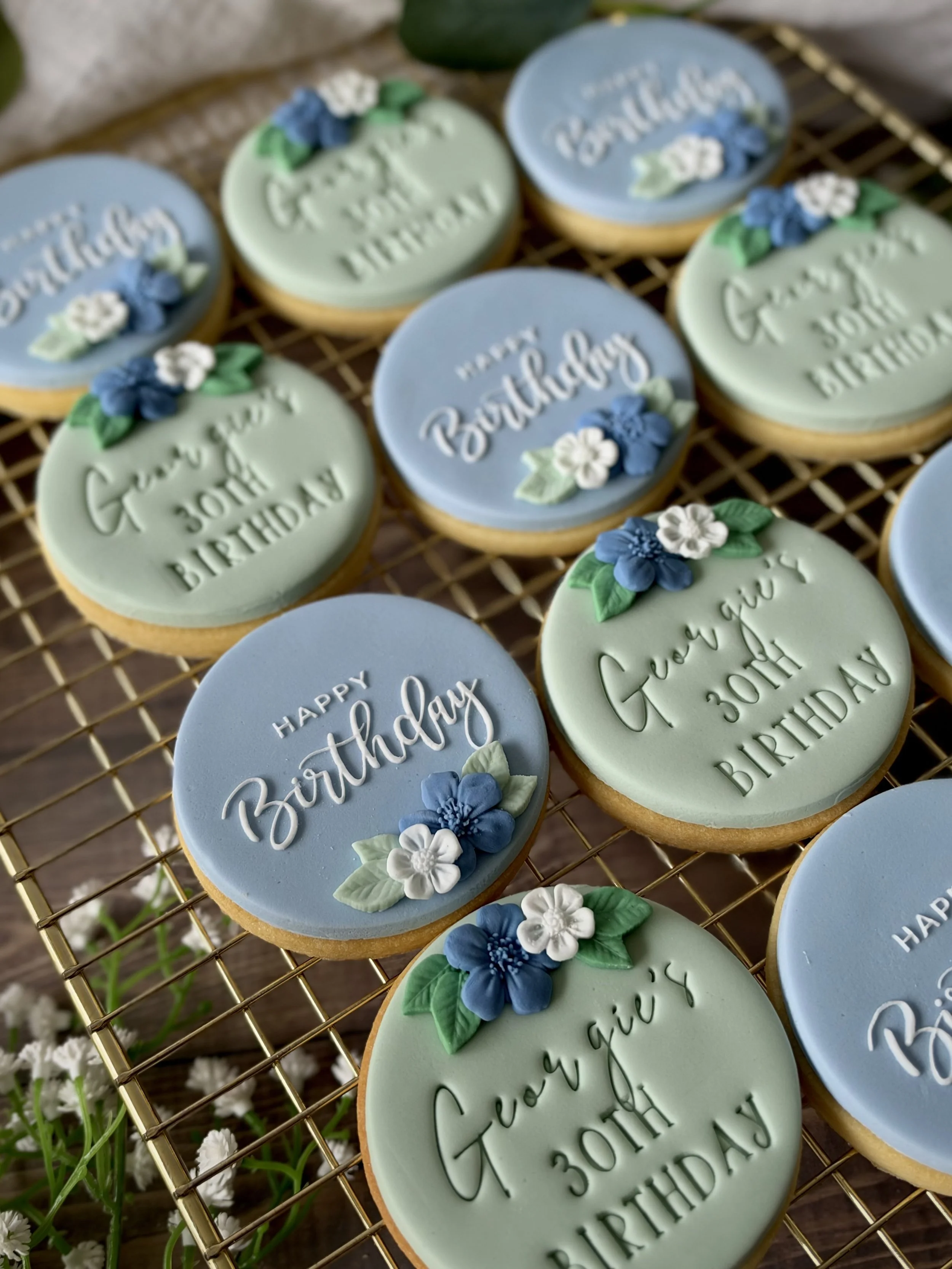 Elegant 30th birthday cookies in soft sage green and dusty blue, featuring personalised lettering and delicate handcrafted fondant flowers in shades of blue and white.
