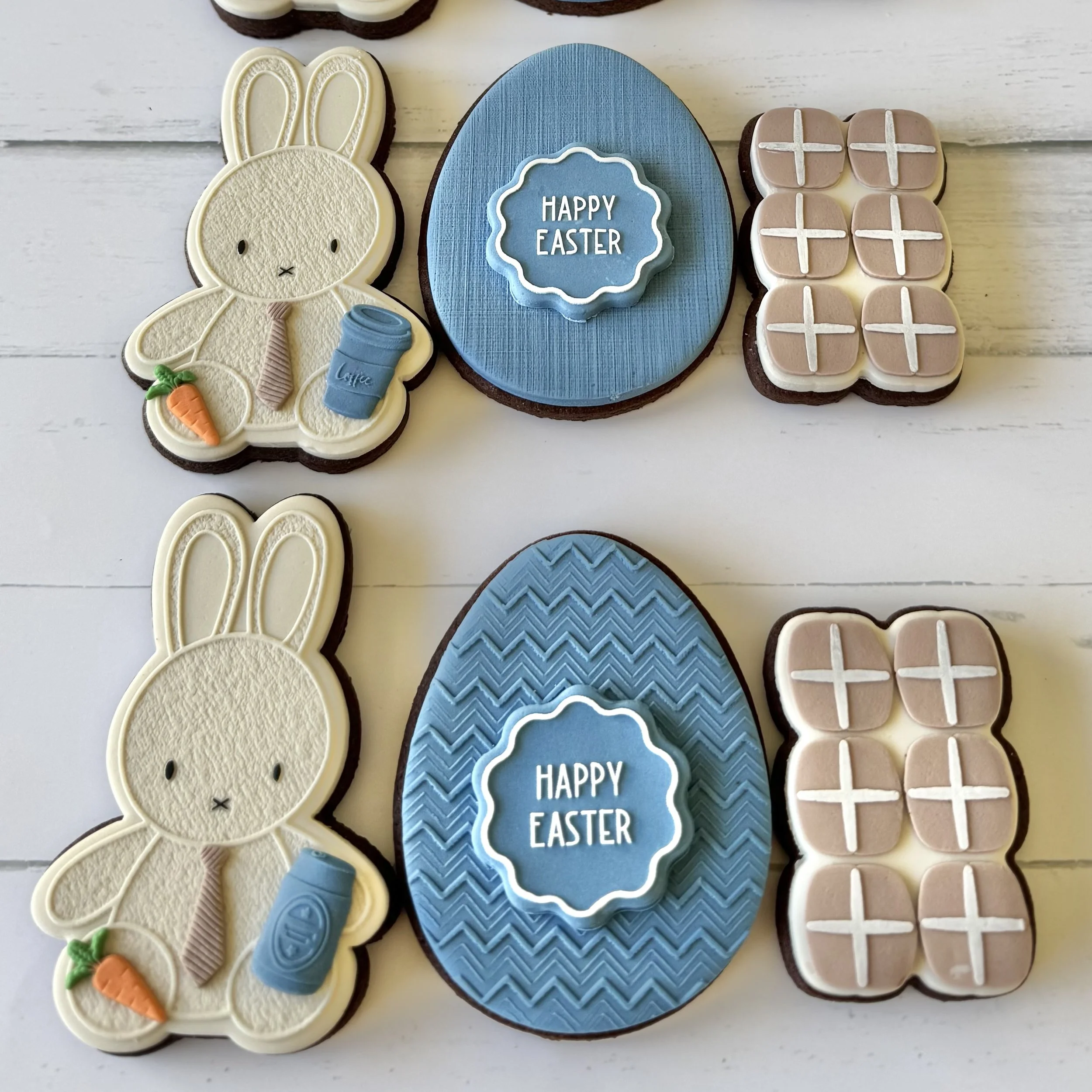 Decorative cookies for Easter, including a bunny with a tie and beer or coffee; a blue egg-shaped cookie with a white border and the message 'Happy Easter'; and a 6-pack of hot cross buns