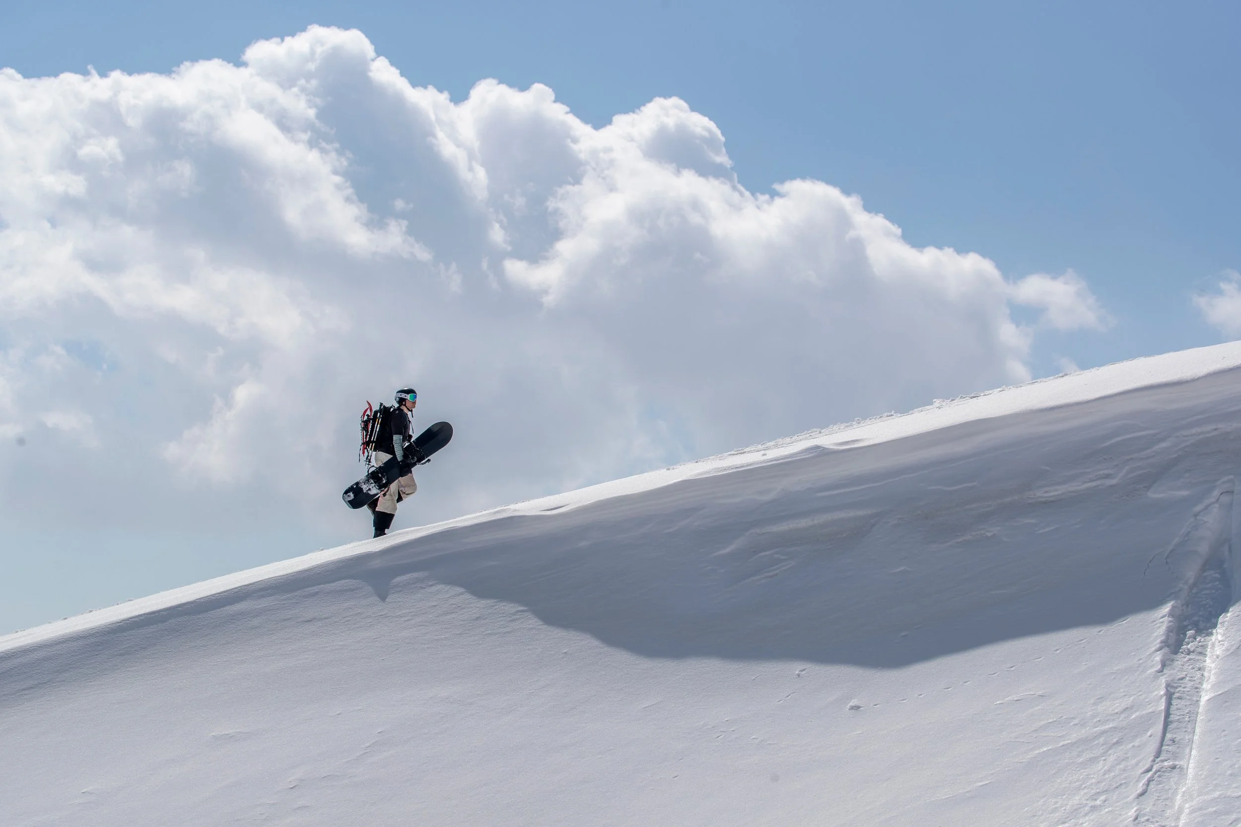 A person dressed in winter gear, including a helmet and goggles, is carrying a snowboard and walking up a snowy slope in a mountain landscape under a partly cloudy blue sky.