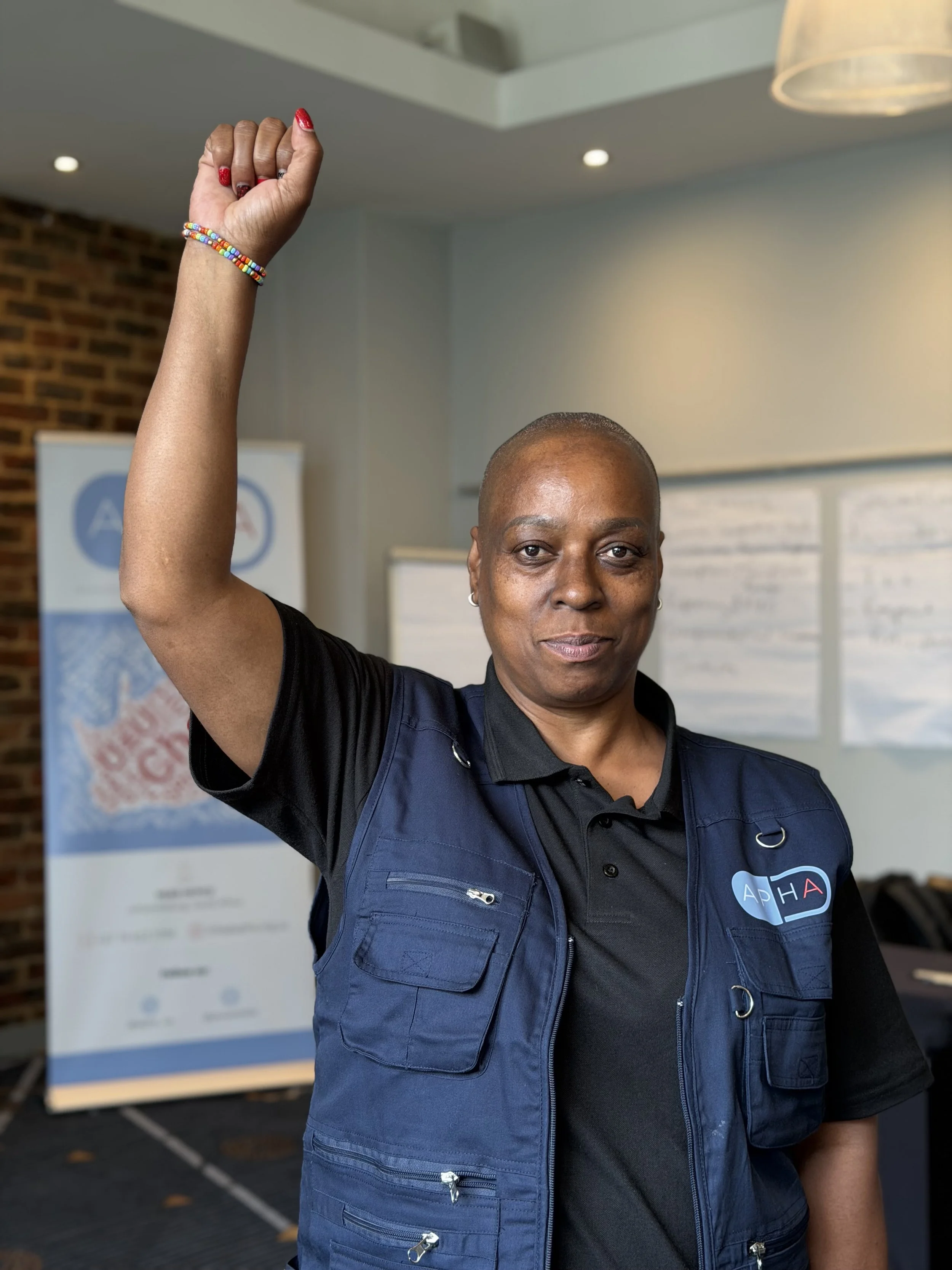 A woman with a shaved head wearing a black shirt and a blue vest that has a logo reading 'APHA.' She is raising her right hand in a fist with red nail polish and colorful beaded bracelet, in an indoor setting with a blurred background and a poster or exhibit.