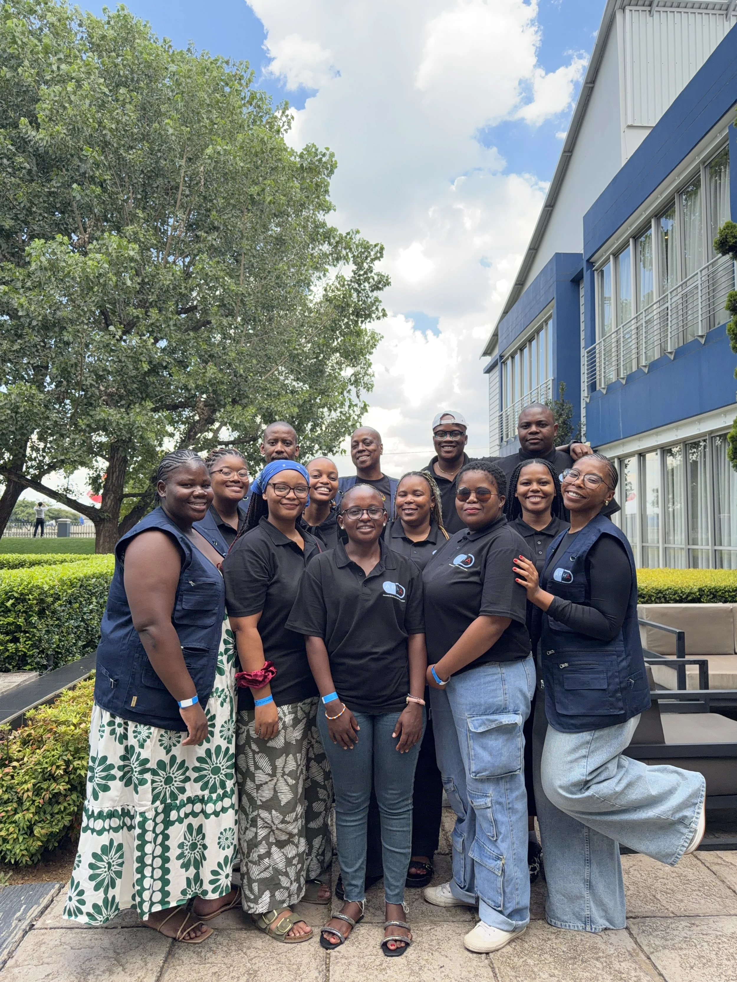 Group of diverse people smiling and posing outdoors in front of a building and trees on a partly cloudy day.