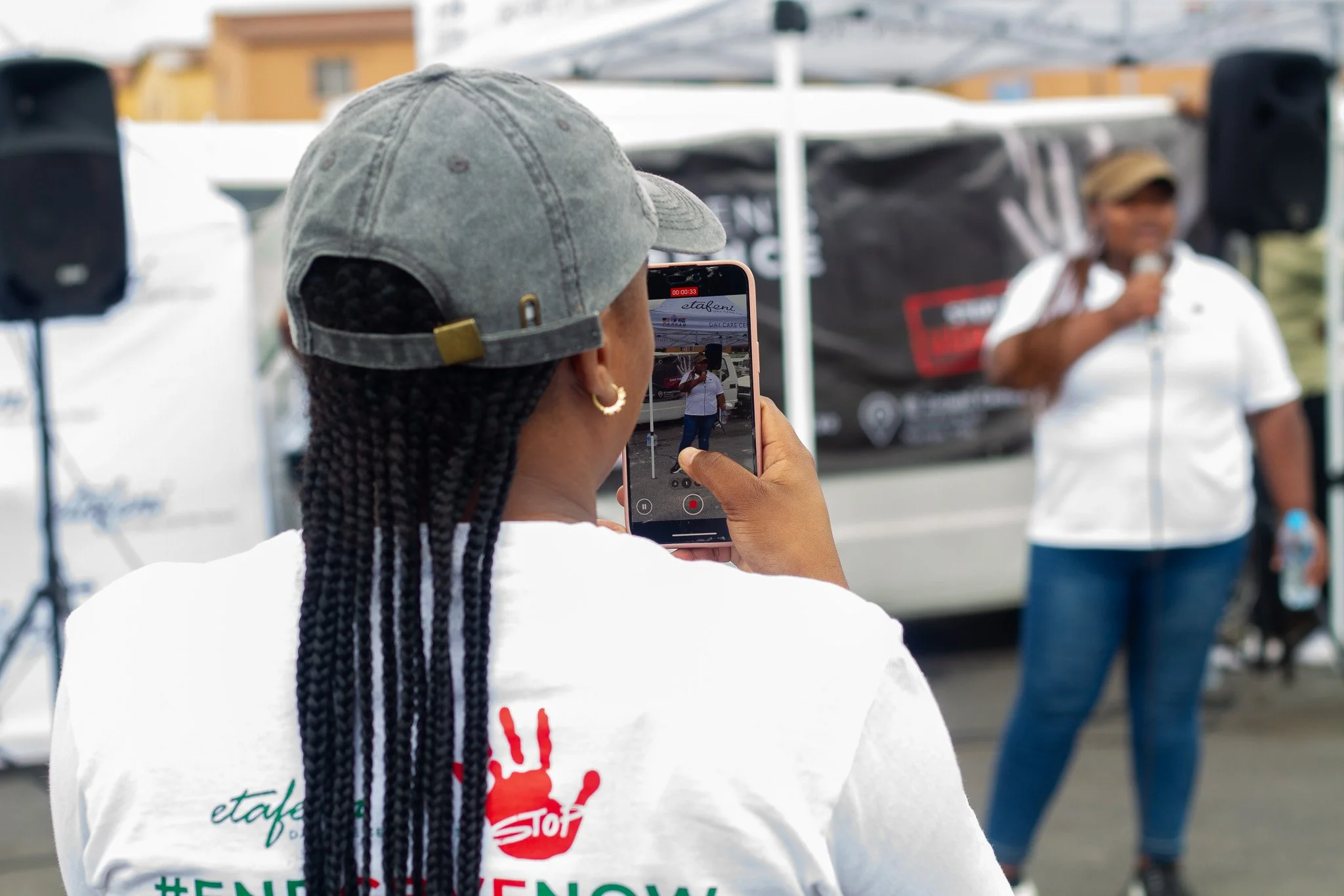 A person with braided hair wearing a gray cap and white shirt taking a photo of a speaker and a woman speaking into a microphone at an outdoor event.