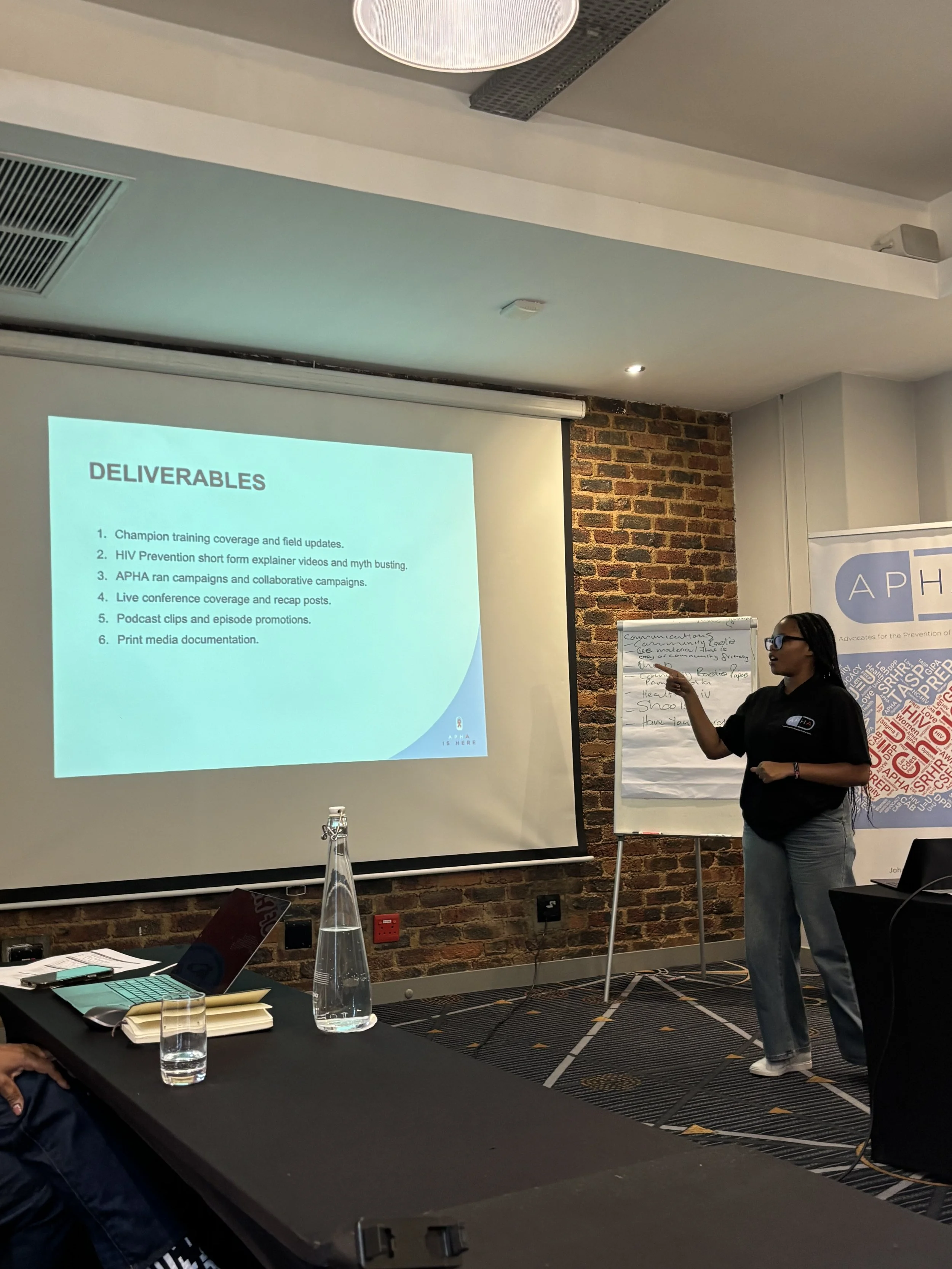 A woman giving a presentation in a conference room with a PowerPoint slide titled 'Deliverables' and a brick wall in the background. There are tables, bottles of water, and laptops visible in the room.
