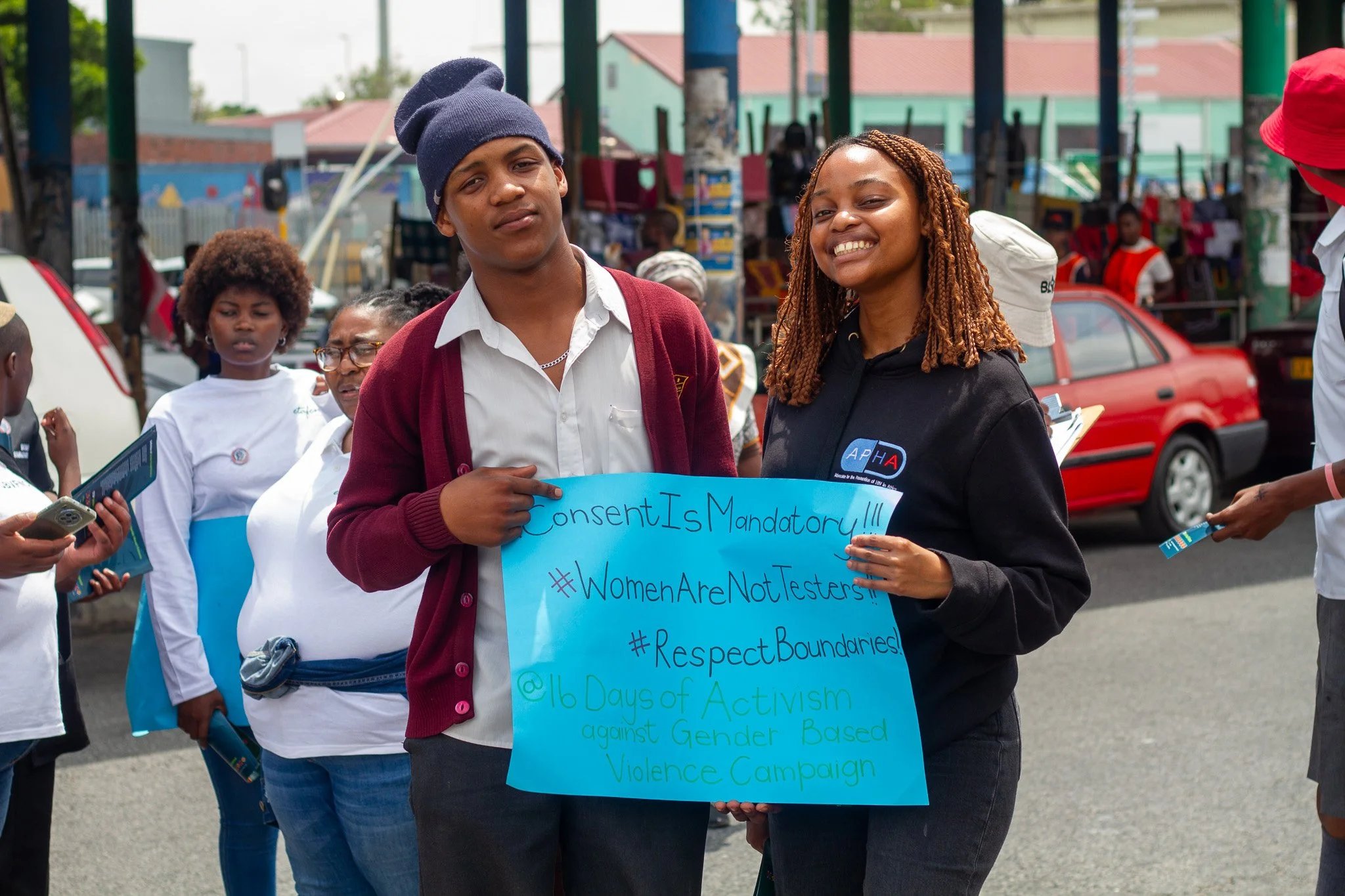 Two young women at a protest holding a blue sign with messages about activism against gender-based violence. They are smiling and standing outdoors with other people and cars in the background.