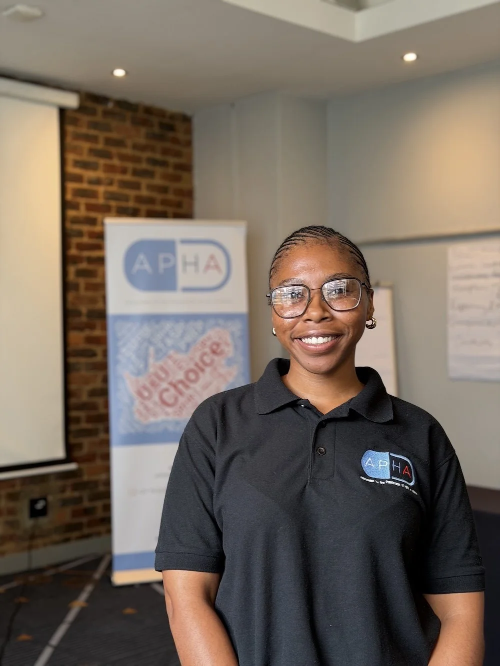 A smiling woman with glasses and braids wearing a black polo shirt with a logo, standing in a conference room with a banner and flipchart in the background.
