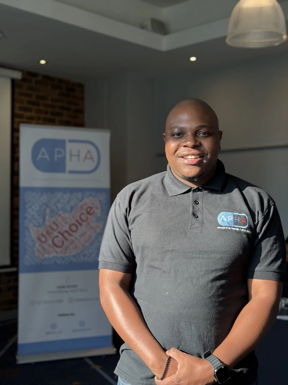 A smiling man wearing a black polo shirt with a logo, standing indoors in front of a banner that reads 'UEFA Choice' and features the APHA logo.