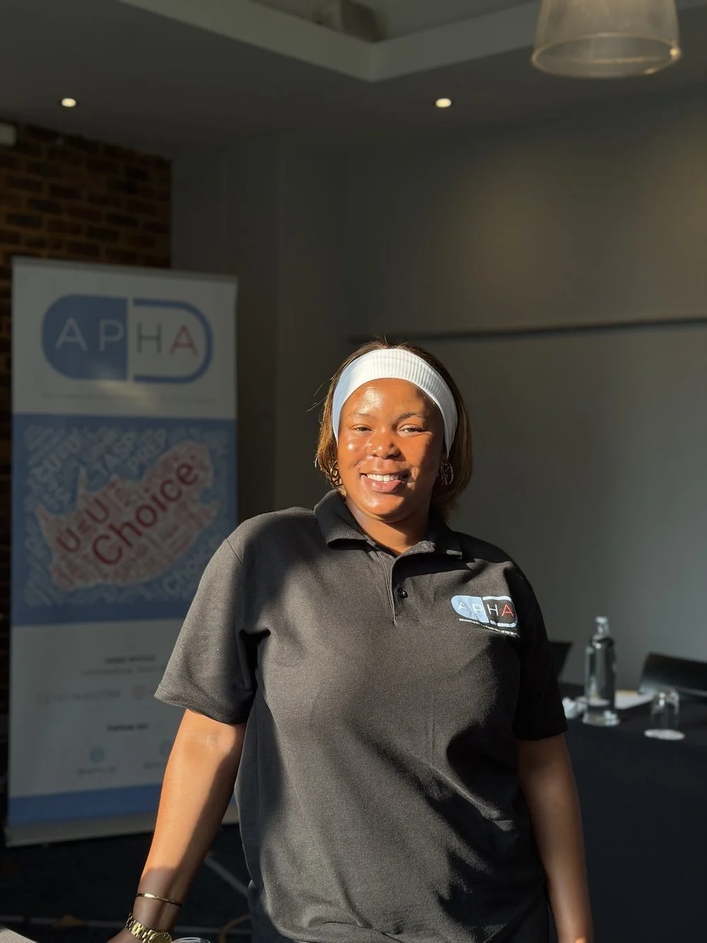 Woman smiling wearing a black polo shirt with a logo, standing indoors with a banner in the background.
