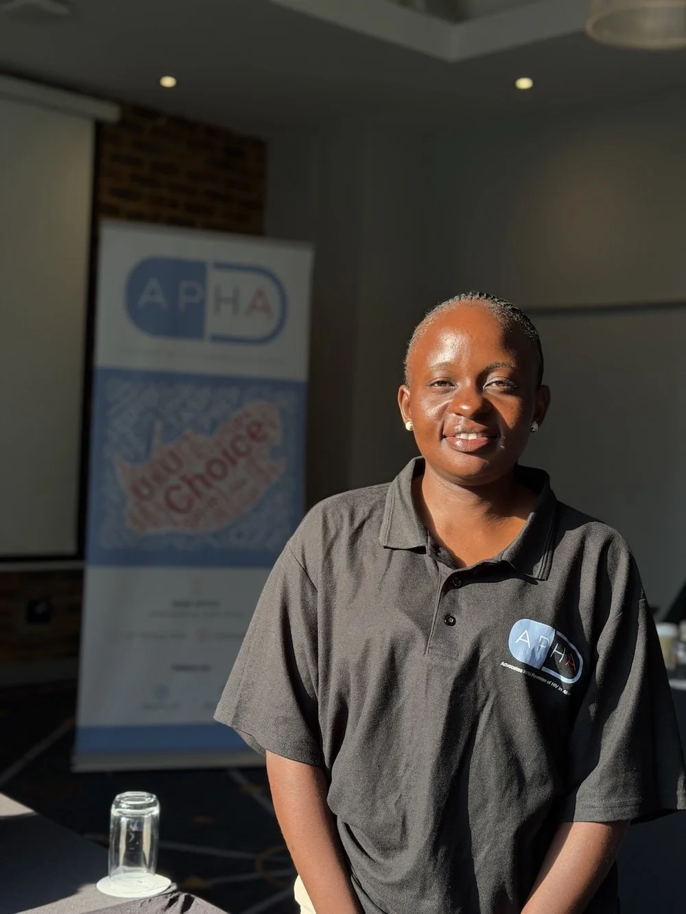 A woman in a gray polo shirt with an APHA logo stands indoors near a table with glasses, smiling at the camera. A banner with APHA branding and the slogan "Making Better Choices" is in the background.