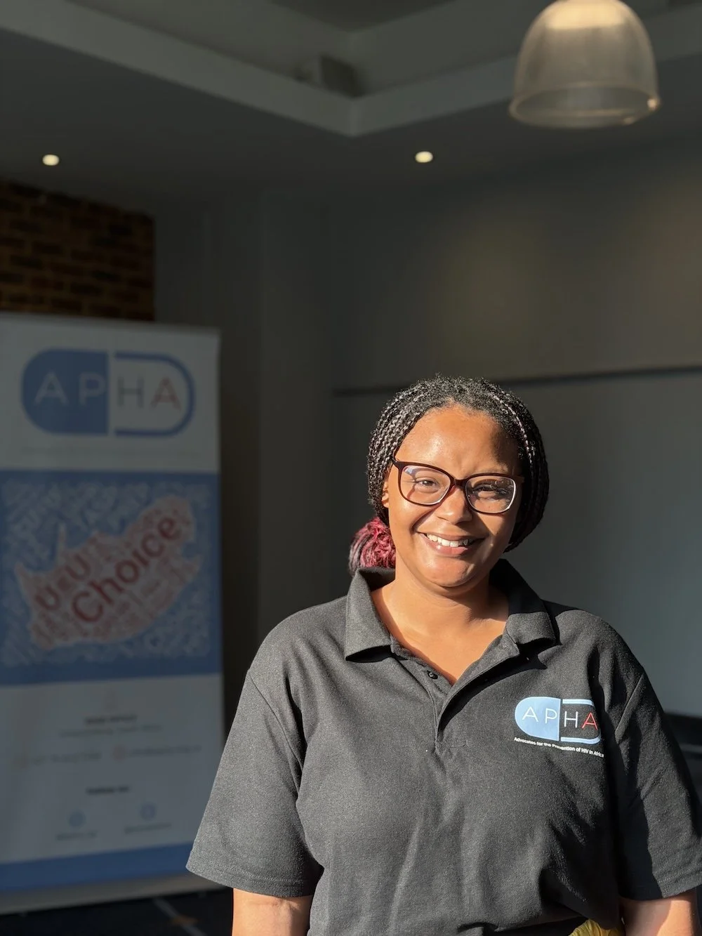 Smiling woman wearing glasses and a gray polo shirt with an 'APHA' logo standing in front of a banner with the words 'HIV Chnage' in a room with ceiling lights.