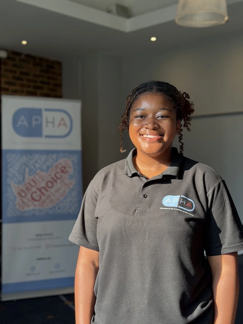 Smiling young woman in a gray polo shirt standing indoors with a poster in the background advocating for HIV prevention.