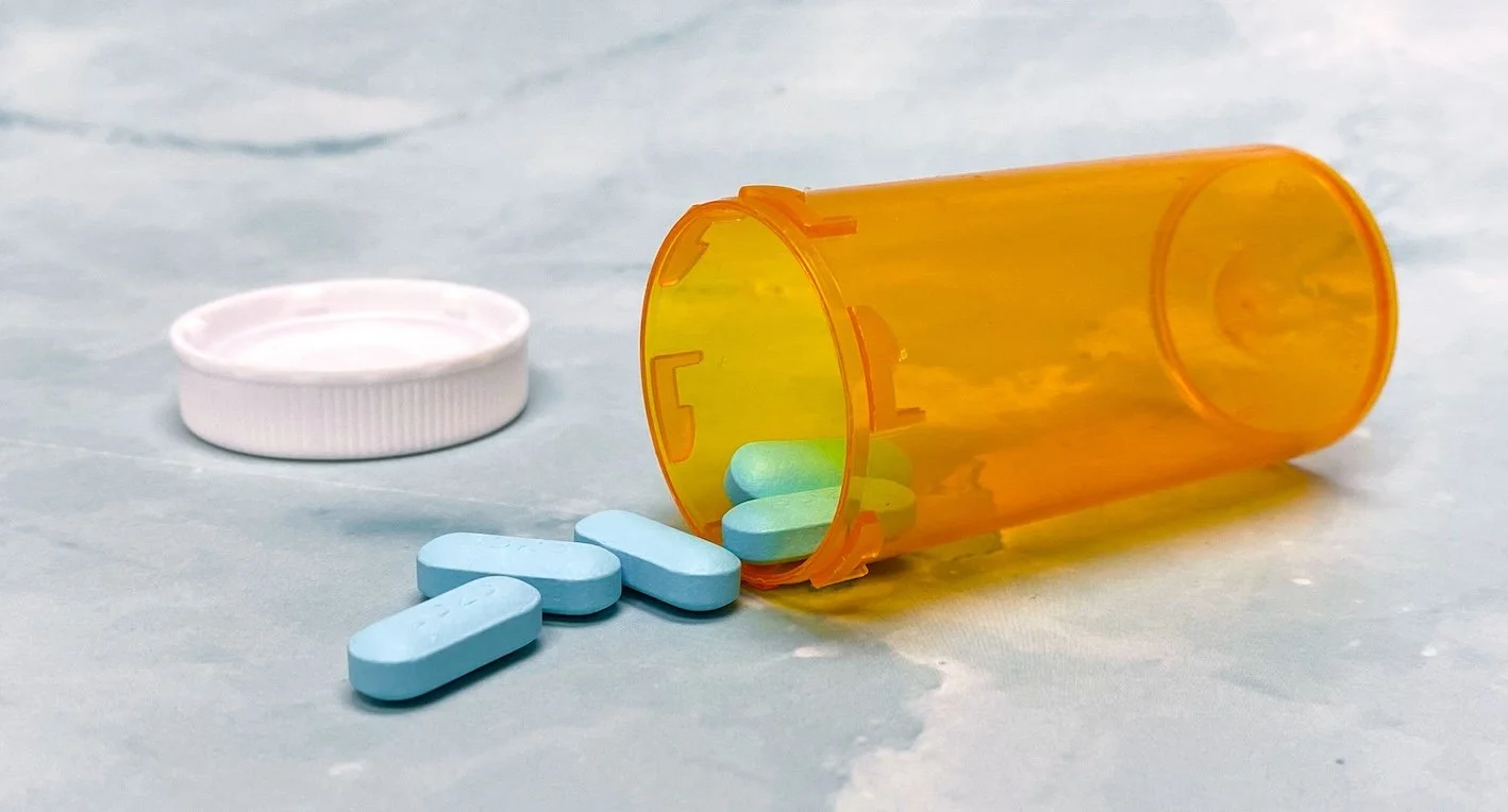 Orange pill bottle with blue pills spilled on light blue surface, white cap beside the bottle.