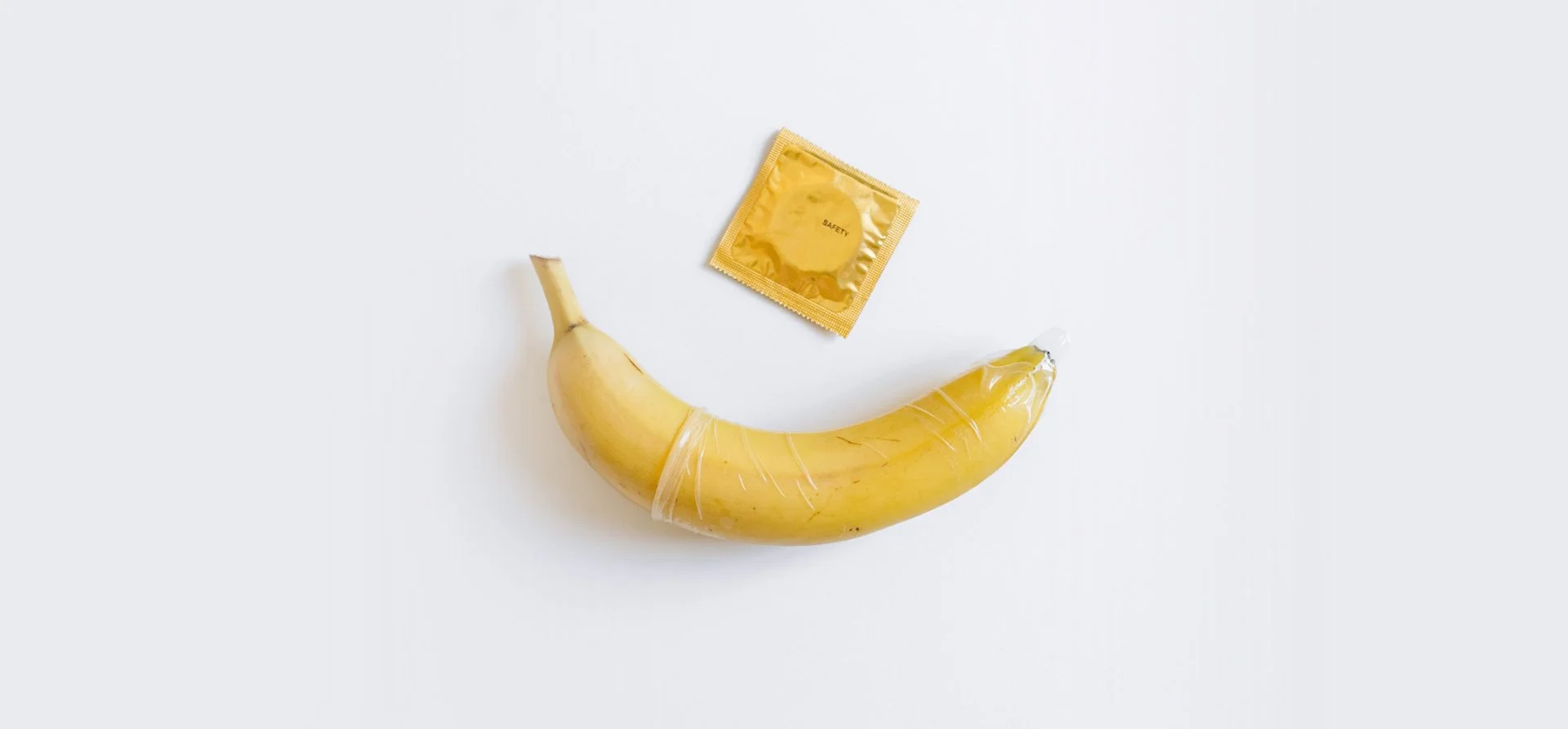A banana with a condom wrapped around it and a gold foil condom wrapper nearby on a white background.