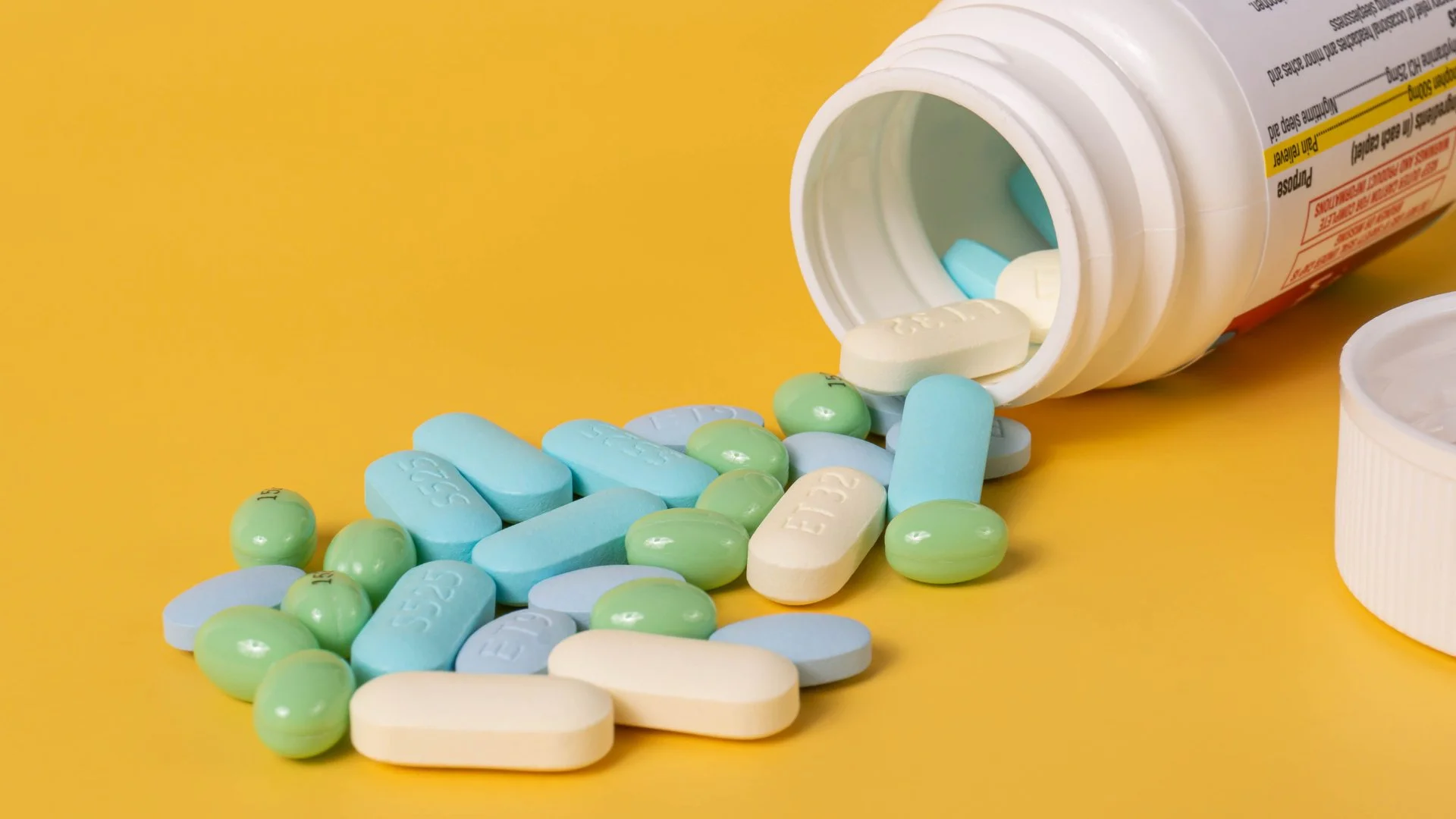A spill of blue, white, and green pills and tablets coming out of a white prescription pill bottle against a yellow background.