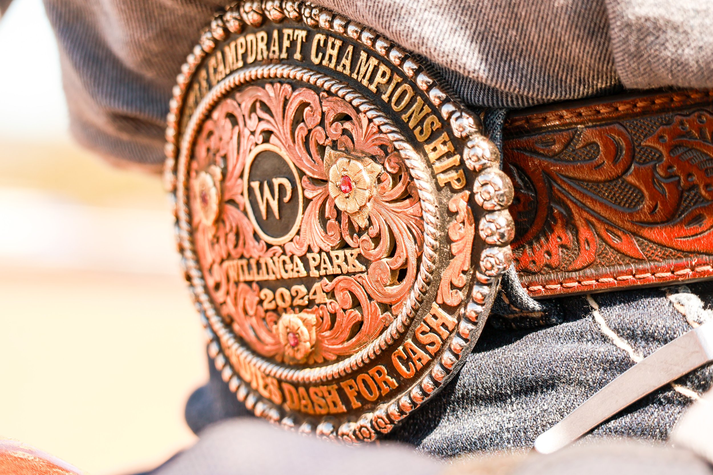 Close-up image of a detailed leather western-style belt buckle with floral and scrollwork designs, and the phrase "WILLINGEA PARK DASH FOR CASH 2024" inscribed around the edge.