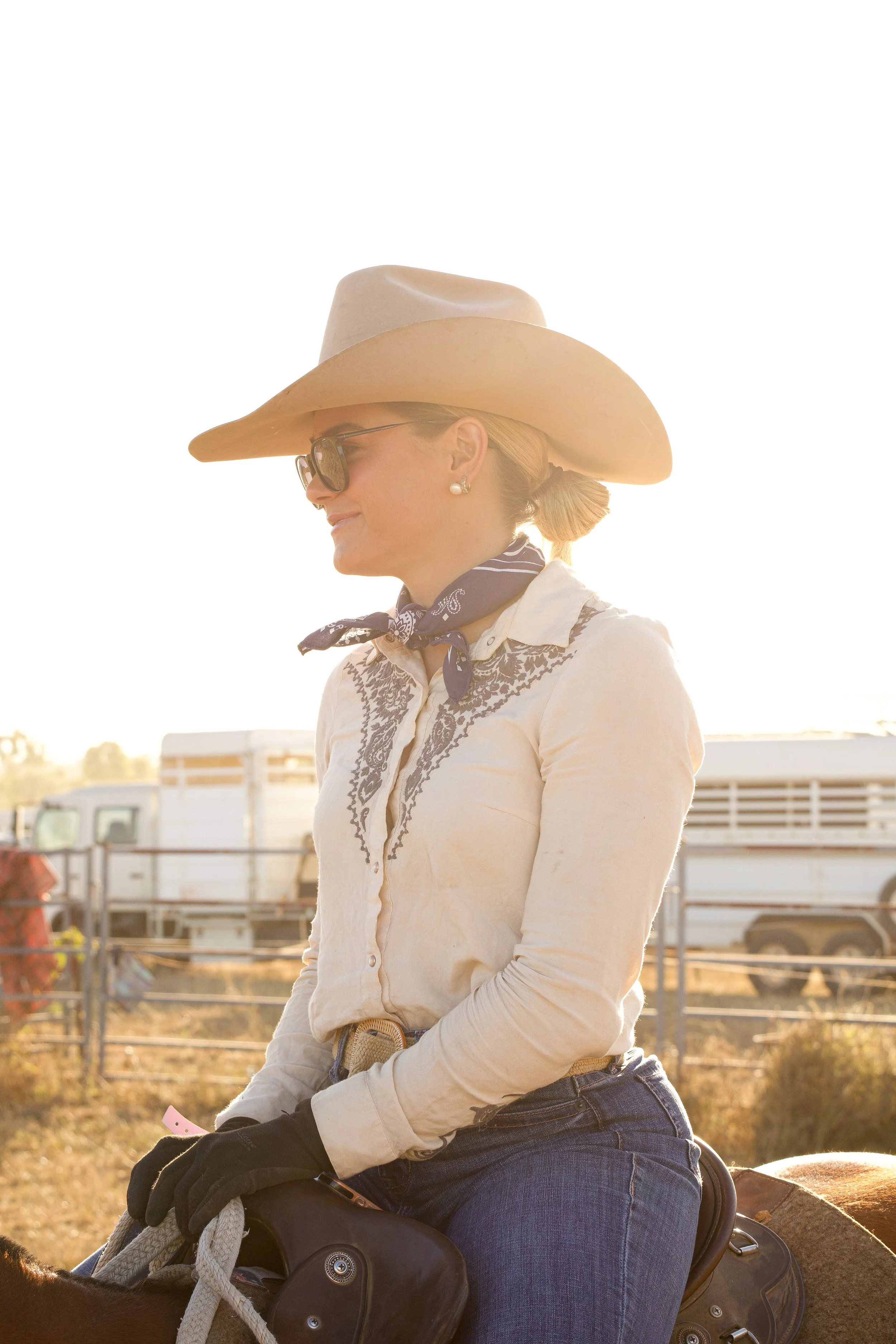 A woman wearing a large cowboy hat, sunglasses, and pearl earrings, smiling while sitting on a horse in a sunny outdoor setting with fences and buildings in the background.