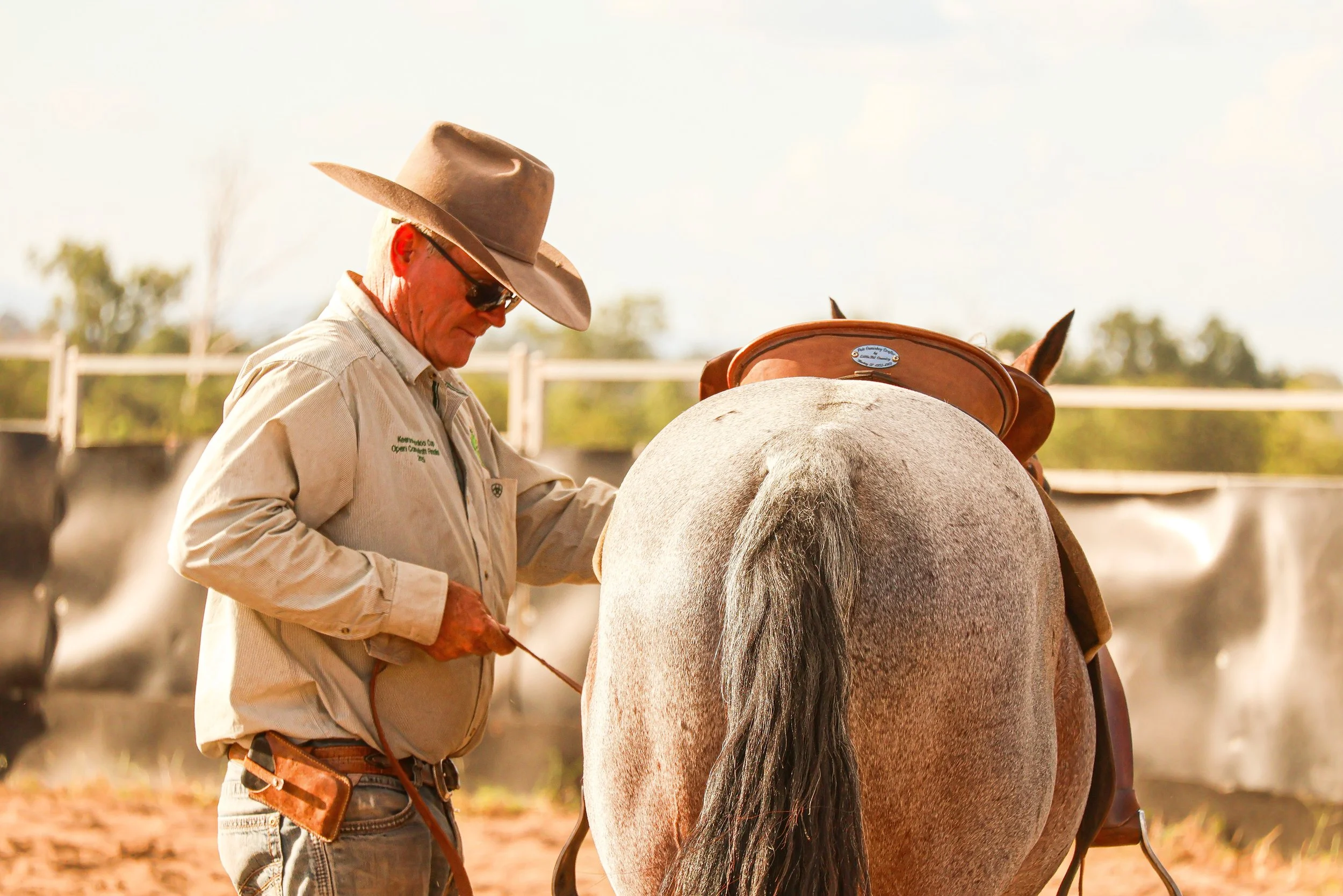 A man wearing a cowboy hat and sunglasses tending to a gray horse in a rural outdoor setting.