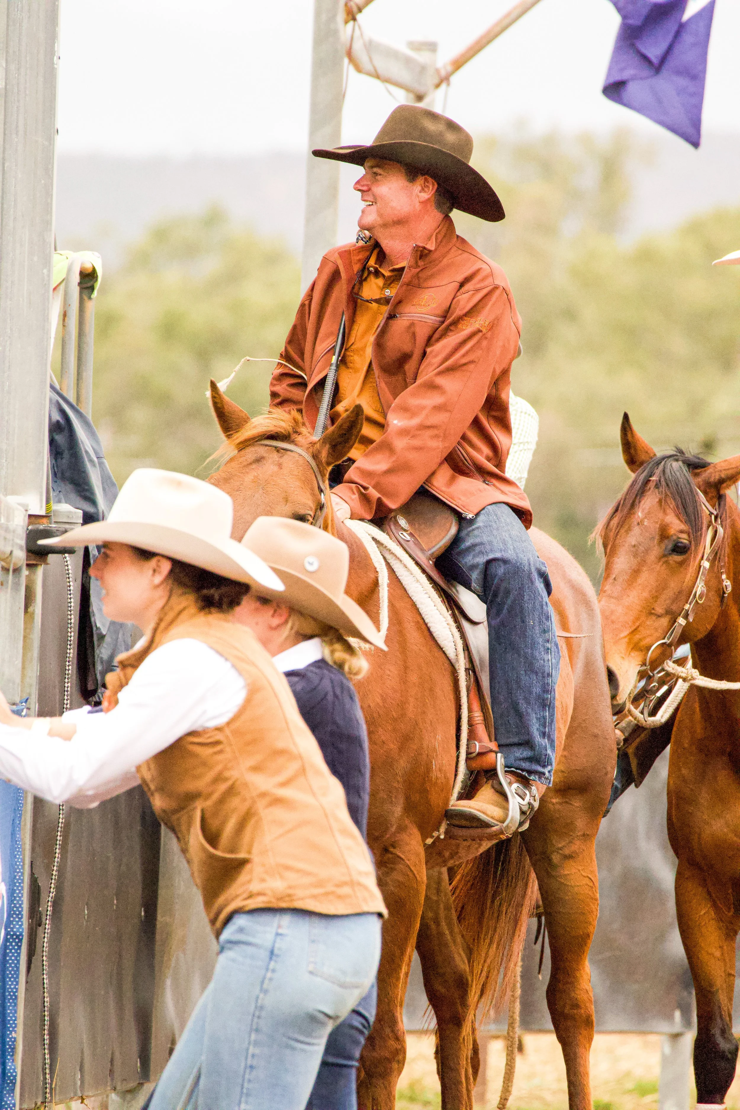 People and horses at a rodeo or livestock event, with a man in a cowboy hat and brown jacket, and a woman in a white cowboy hat and vest, standing near a fence.