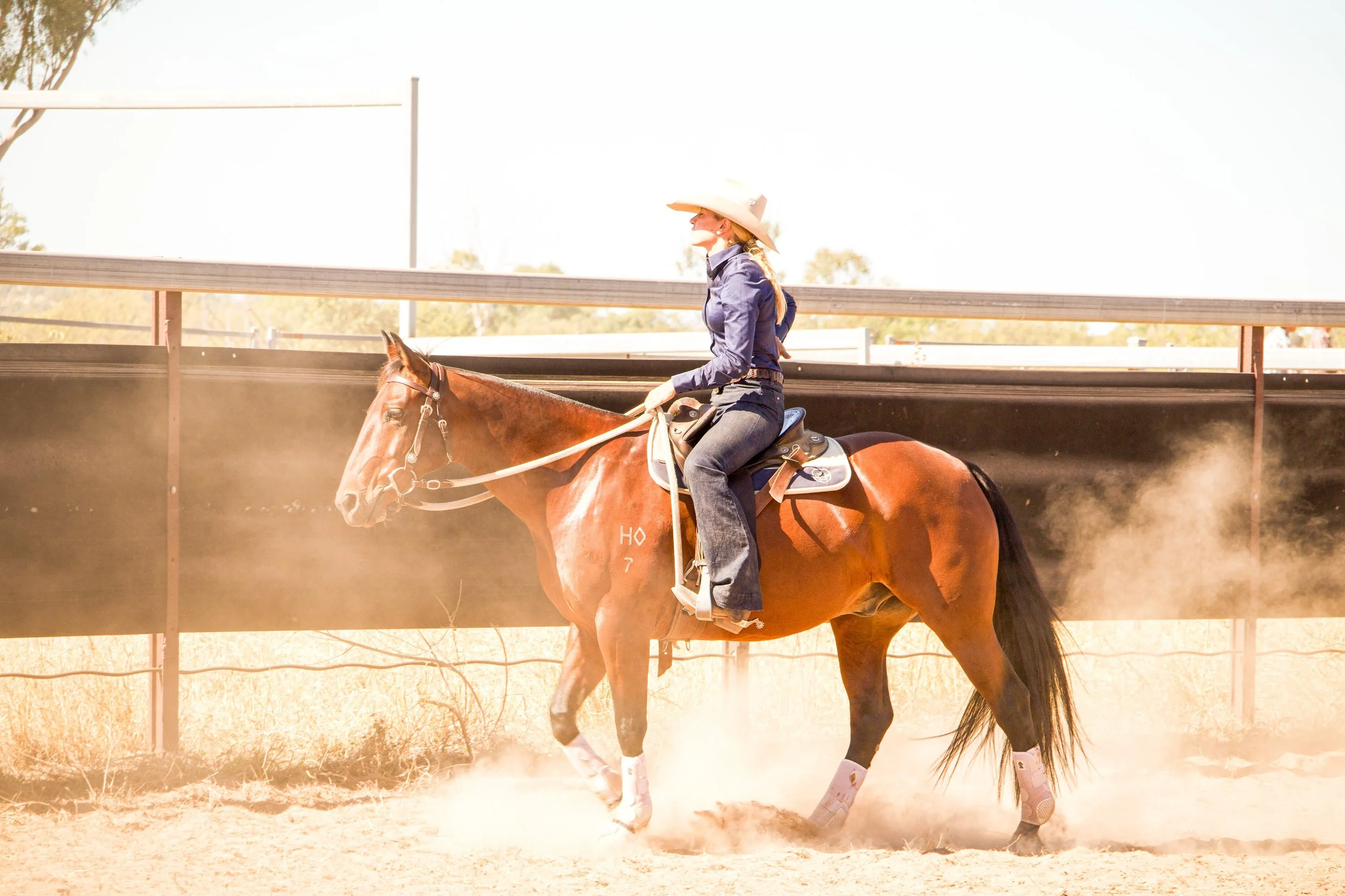 A woman in a cowboy hat and blue shirt riding a horse on a dirt track at a rodeo arena.