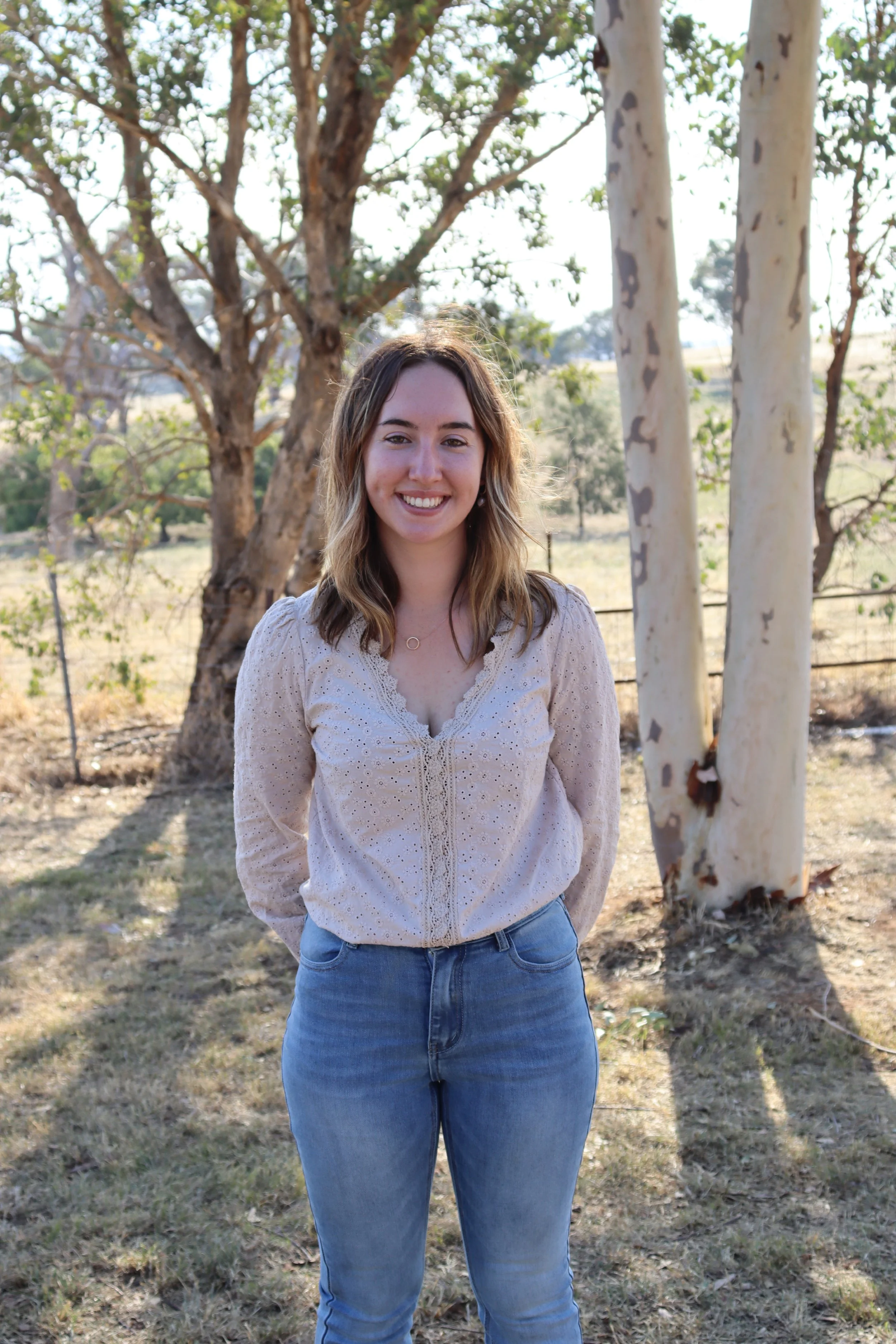 A young woman with shoulder-length light brown hair and fair skin is standing outdoors in front of trees and a landscape during daytime. She is smiling and wearing a beige, embroidered blouse and blue jeans.