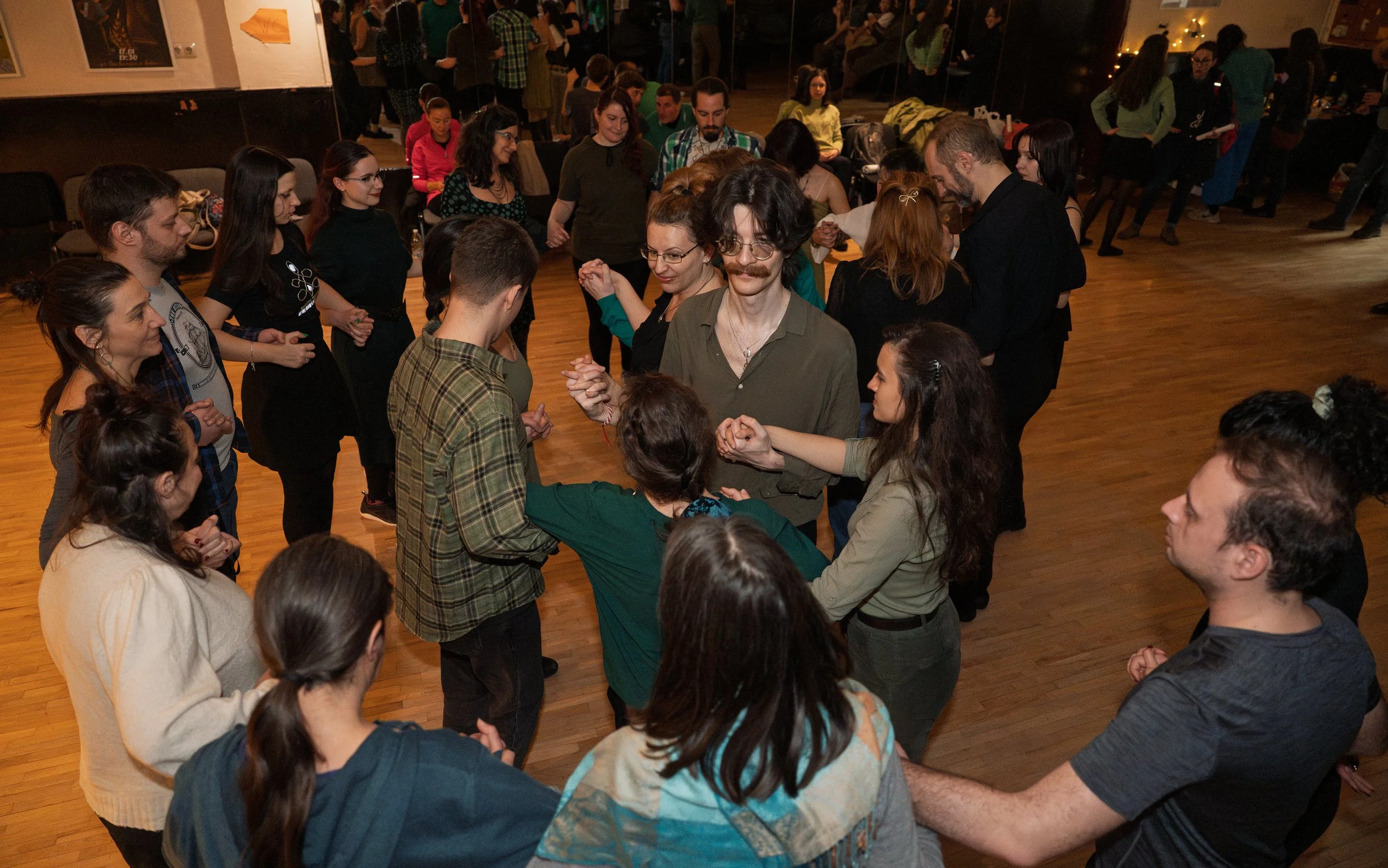 A group of people dancing in a circle in a room with wooden floors, with some others watching or mingling in the background.