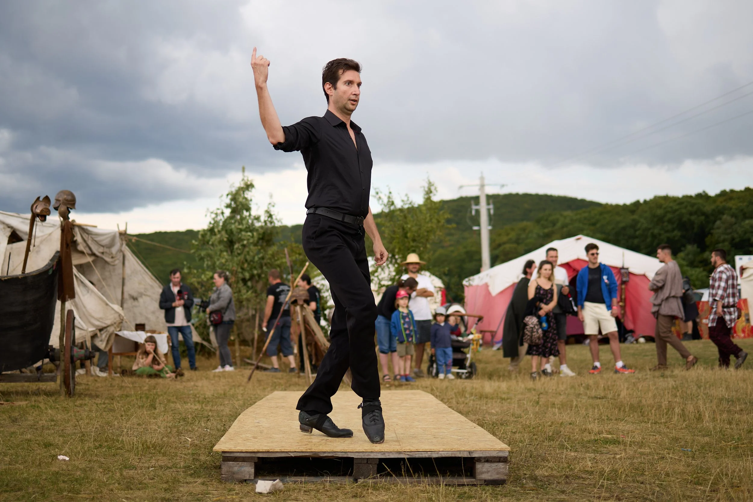 A man in a black shirt and pants on roller skates demonstrates dance moves on a wooden platform at an outdoor event, with tents and a crowd in the background under a cloudy sky.