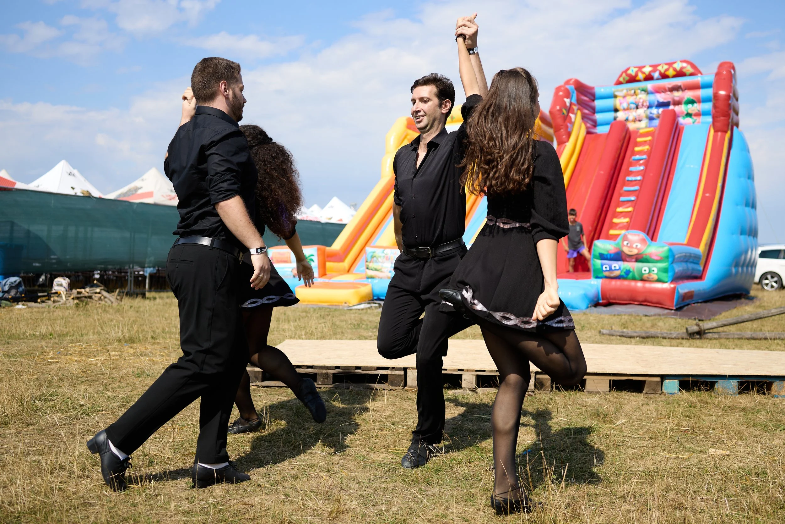 A group of four people dancing outdoors in front of a colorful inflatable slide at a fair or festival on a sunny day.