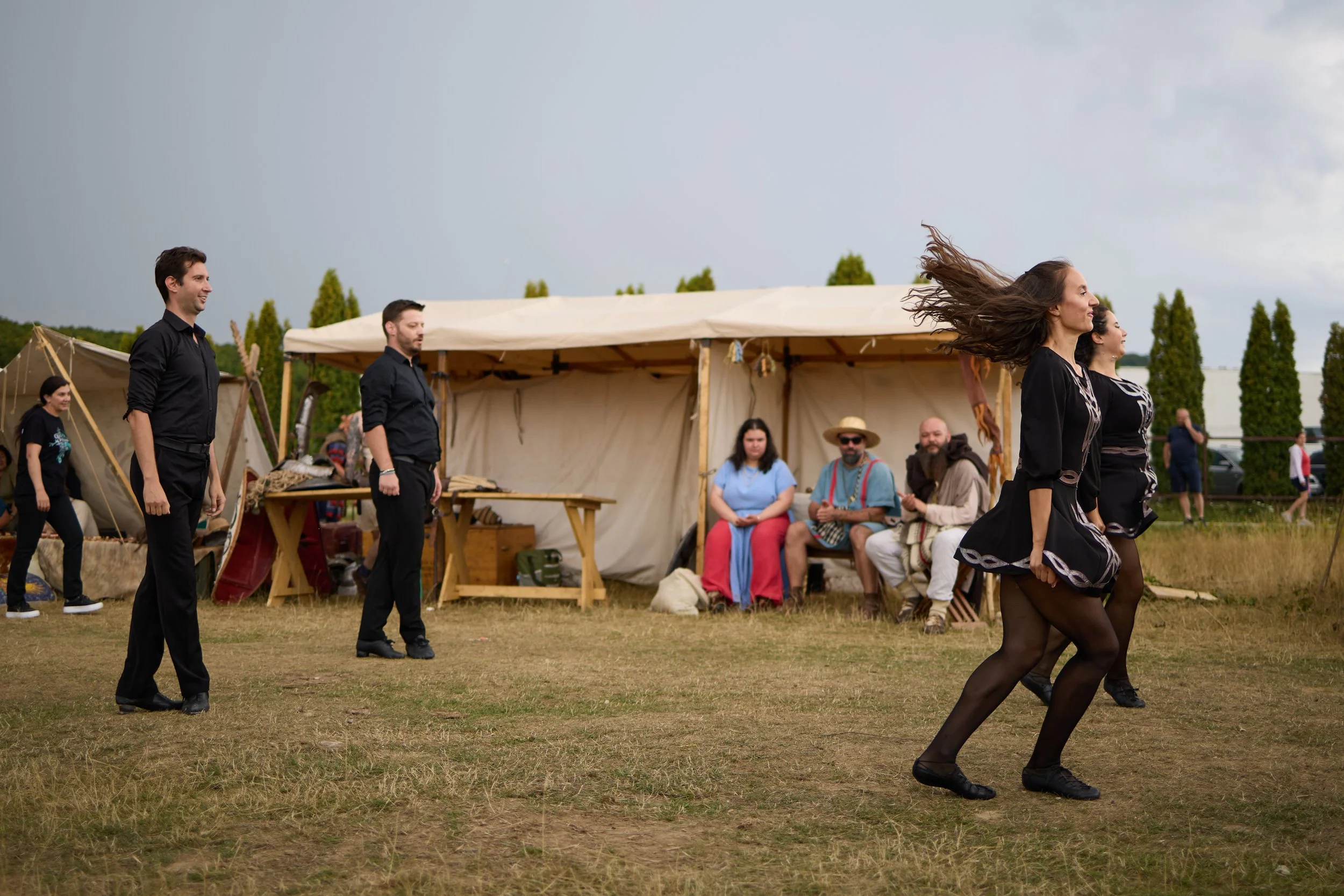 People participating in a traditional dance outdoors during a cultural event, with a tent and onlookers in the background, under cloudy skies.