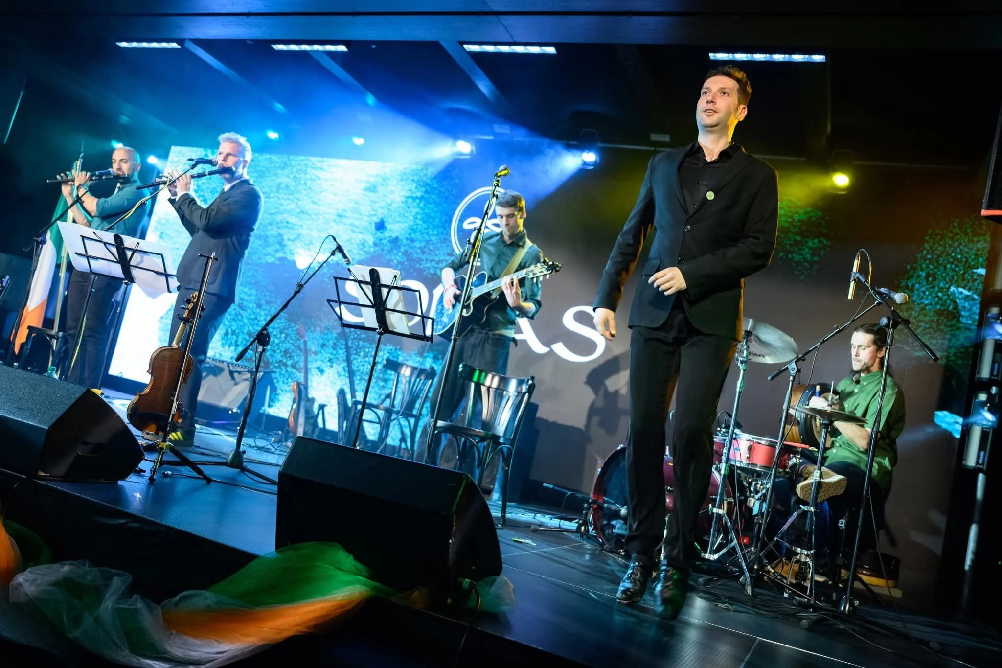 A musical band performing on stage with four musicians playing instruments and a singer. The stage is lit with blue, yellow, and white lights, and there's a large screen behind them displaying abstract visuals and the word 'EAS'.