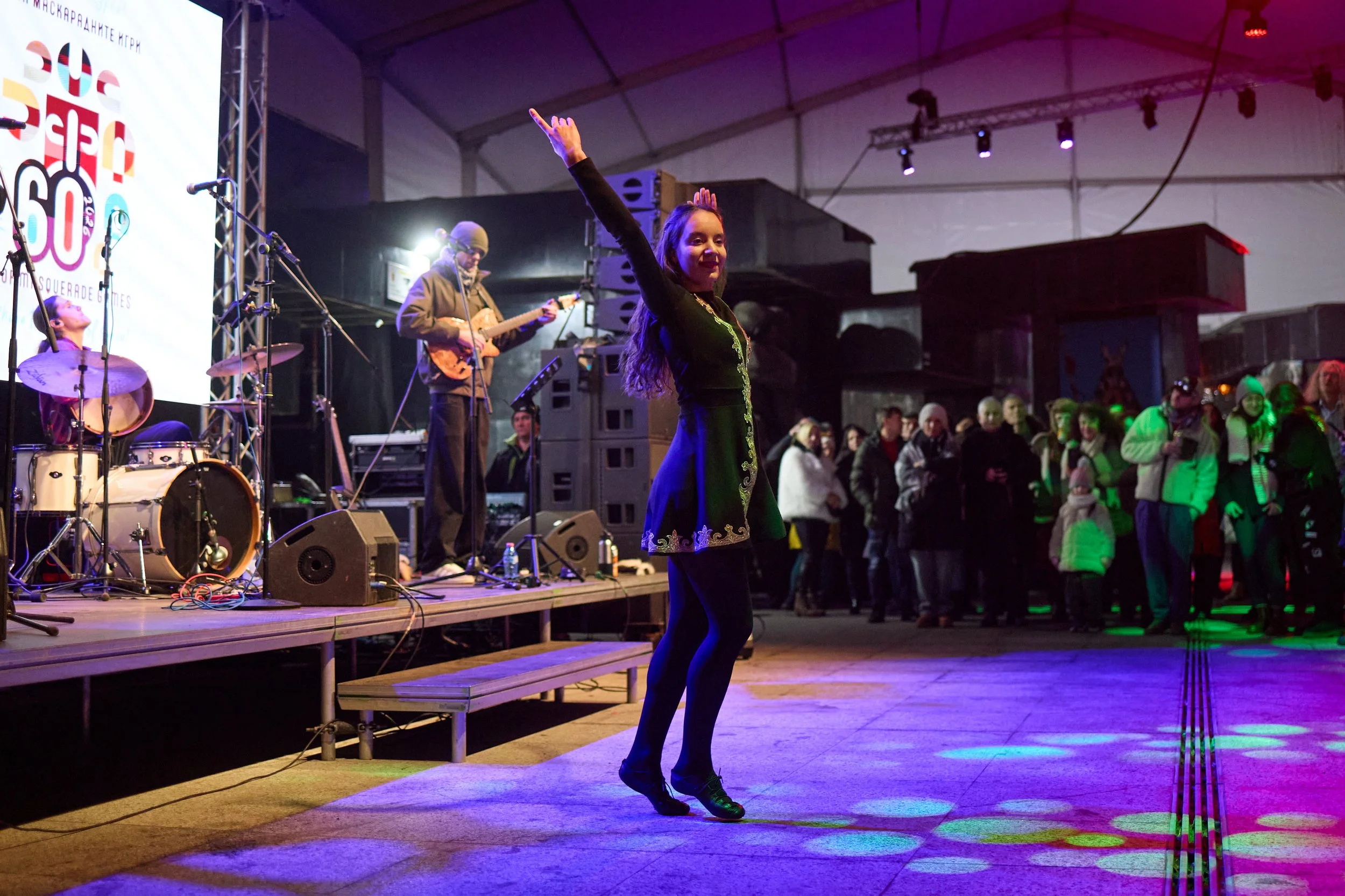 A woman in a black dress with gold embroidery is dancing on a stage at a concert, with an audience in the background and a band playing instruments behind her.