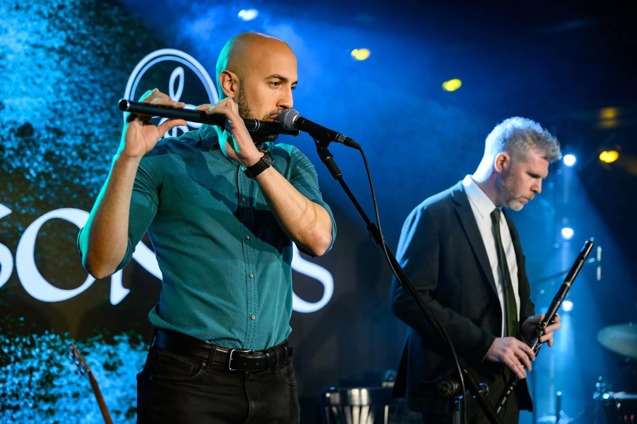 Two men are playing wind instruments on stage under blue lighting, with one man playing a flute and the other playing a saxophone, both dressed in formal attire.