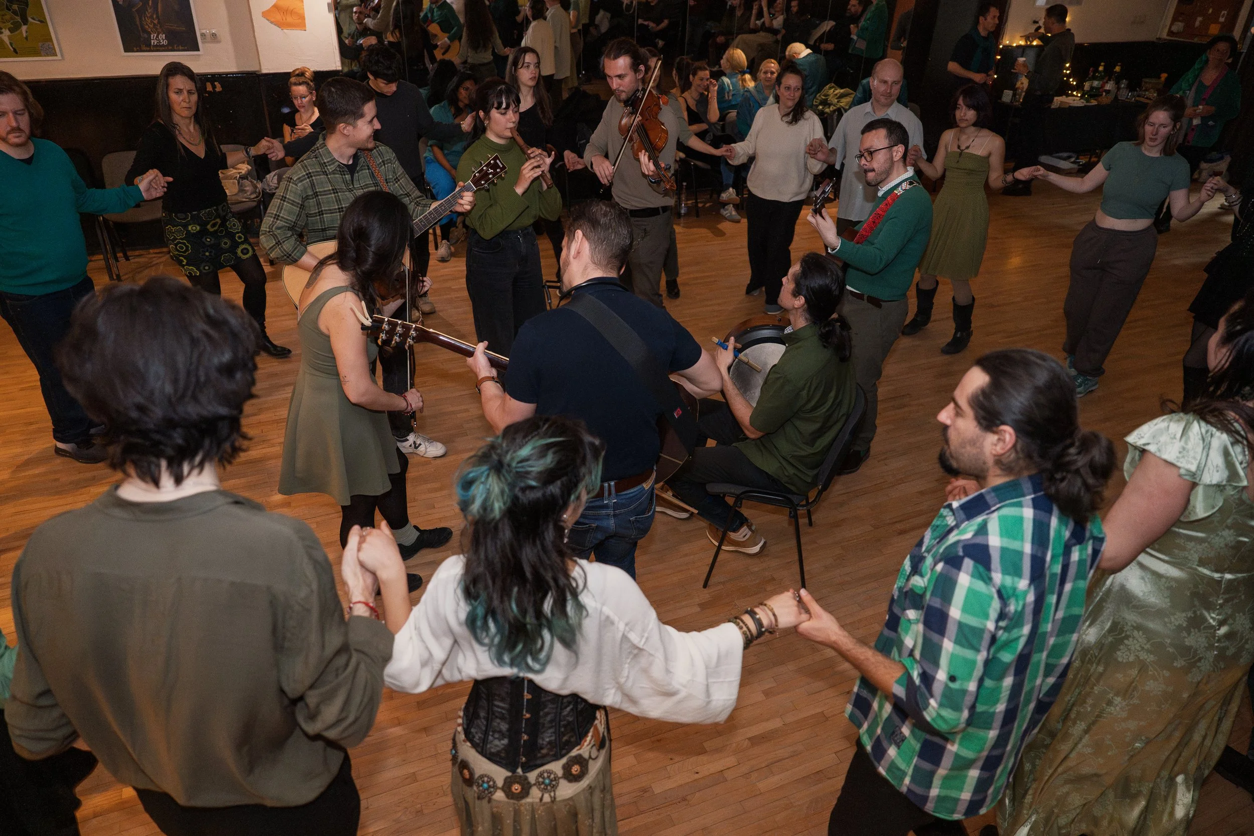 A group of people holding hands in a circle, dancing and playing musical instruments, at a lively indoor gathering or celebration with a wooden floor and some chairs and tables in the background.
