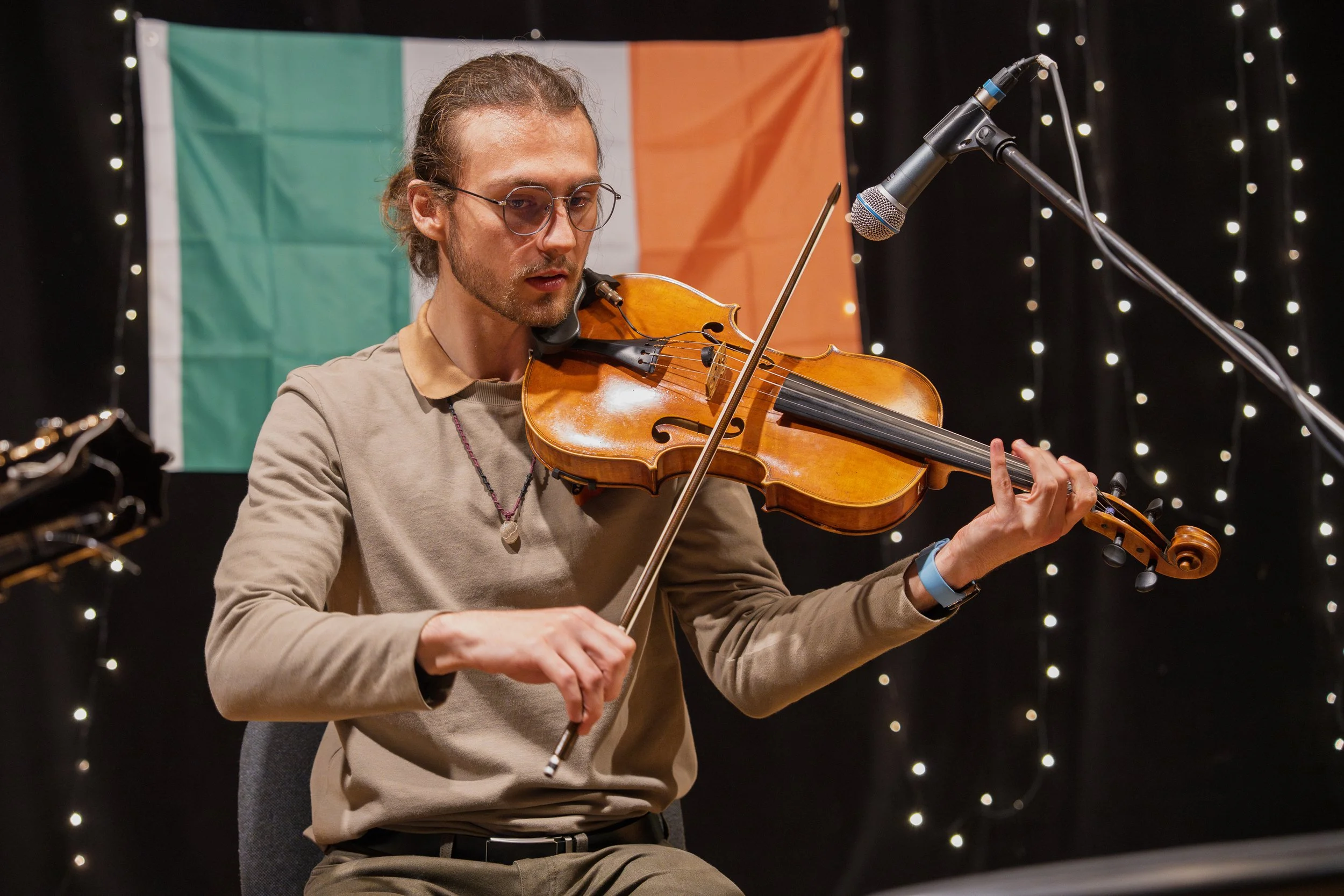 A man with glasses playing the violin in front of an Irish flag with string lights in the background.