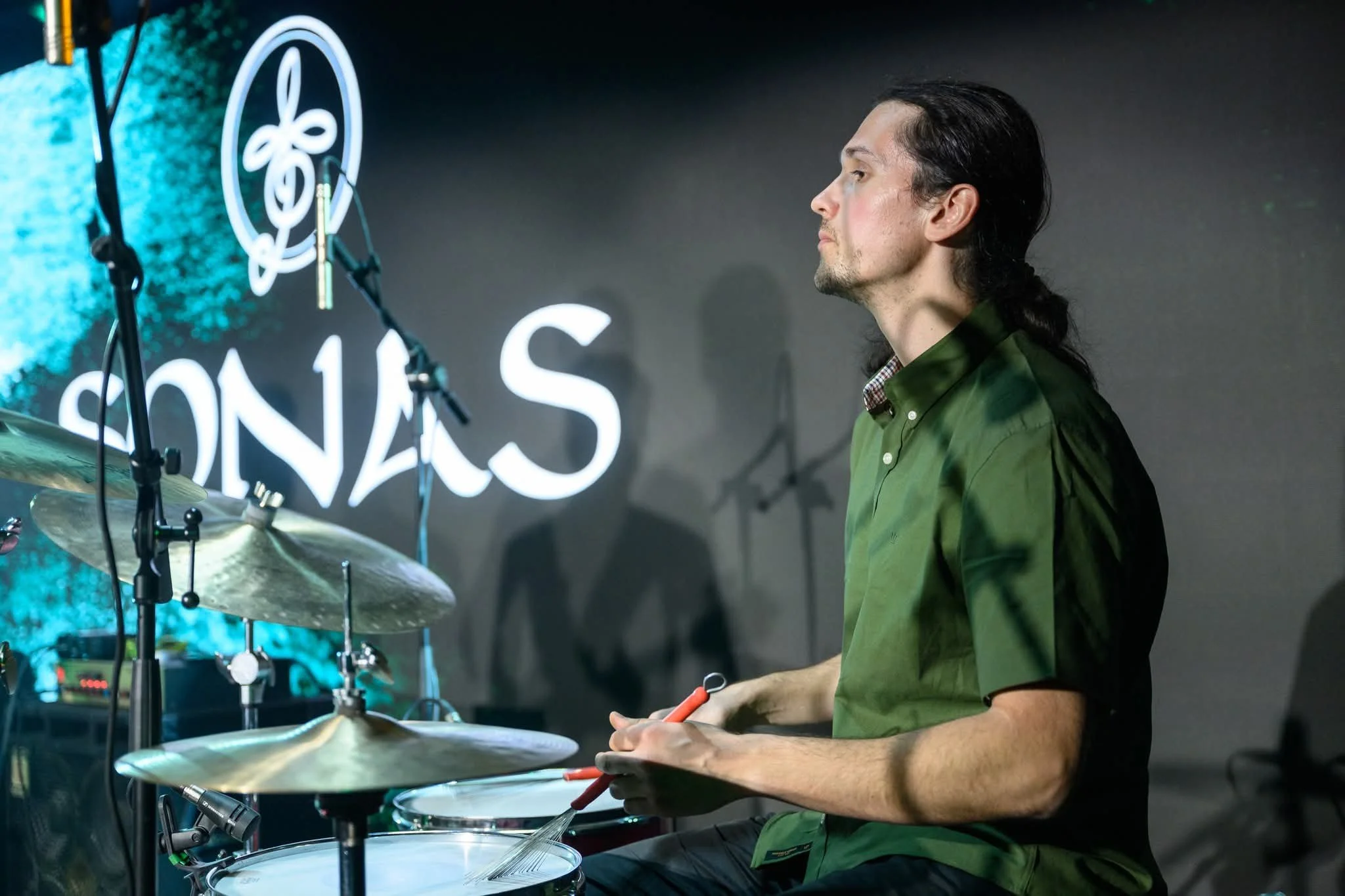 A man with long dark hair, wearing a green shirt, is playing drums with a drumstick in his hand. The drum set is visible with cymbals, and there is a neon sign with a musical note and the word 'SONIC' in the background.