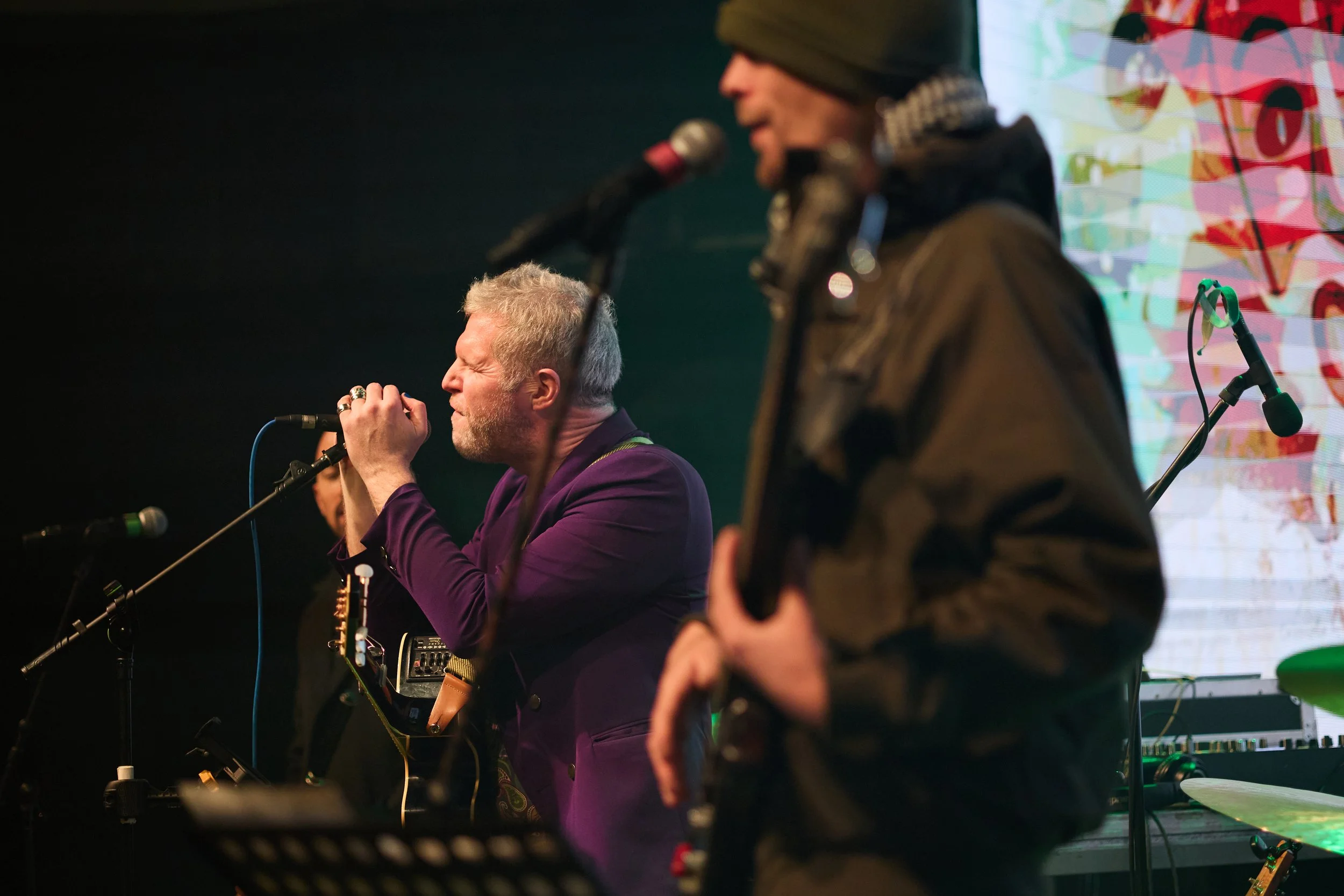 A person singing into a microphone on stage with a colorful backdrop, another person wearing a beanie in the foreground with musical instruments around.