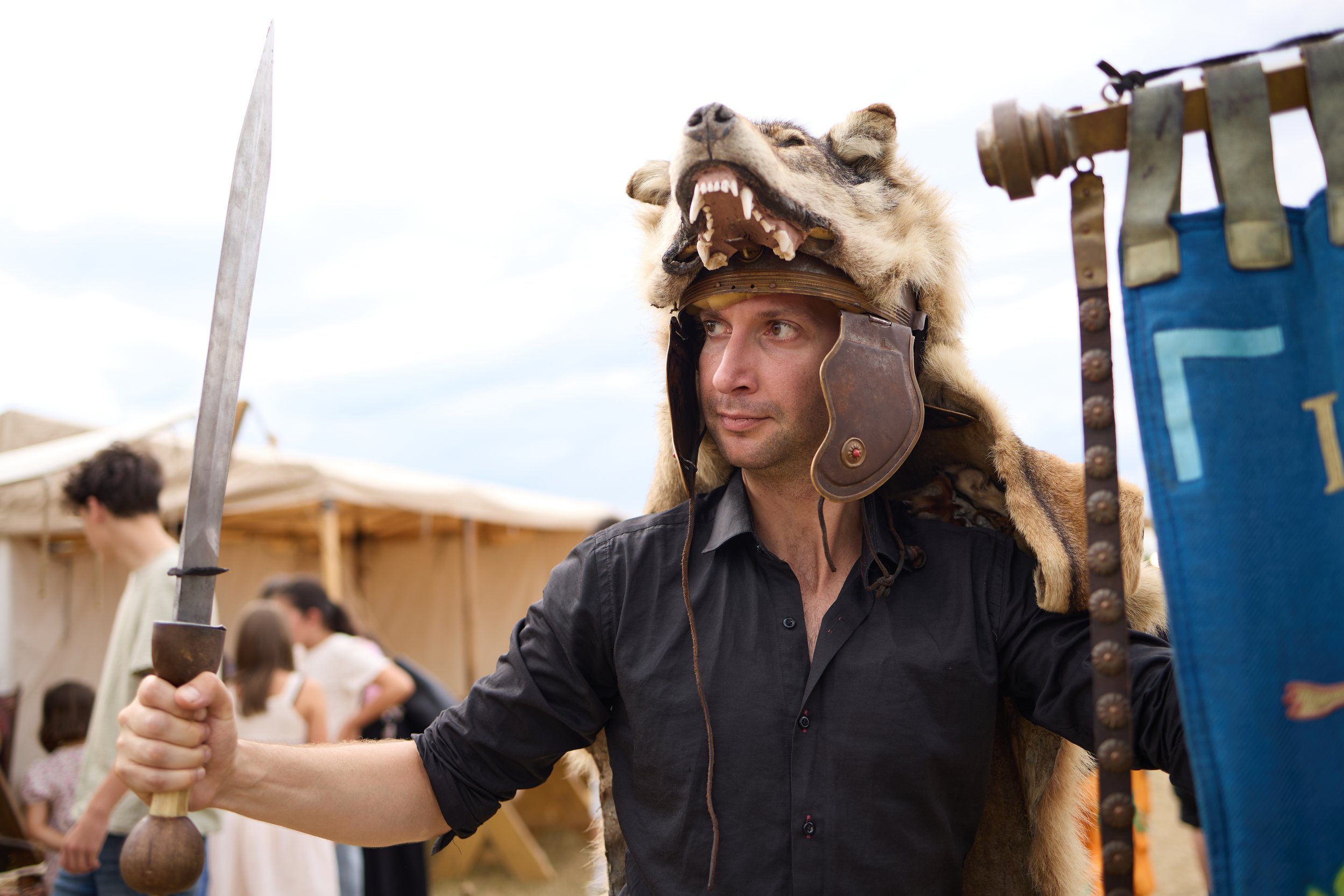 A man dressed in medieval armor wearing a wolf headdress during an outdoor event, holding a long wooden stick, with people and tents visible in the background.