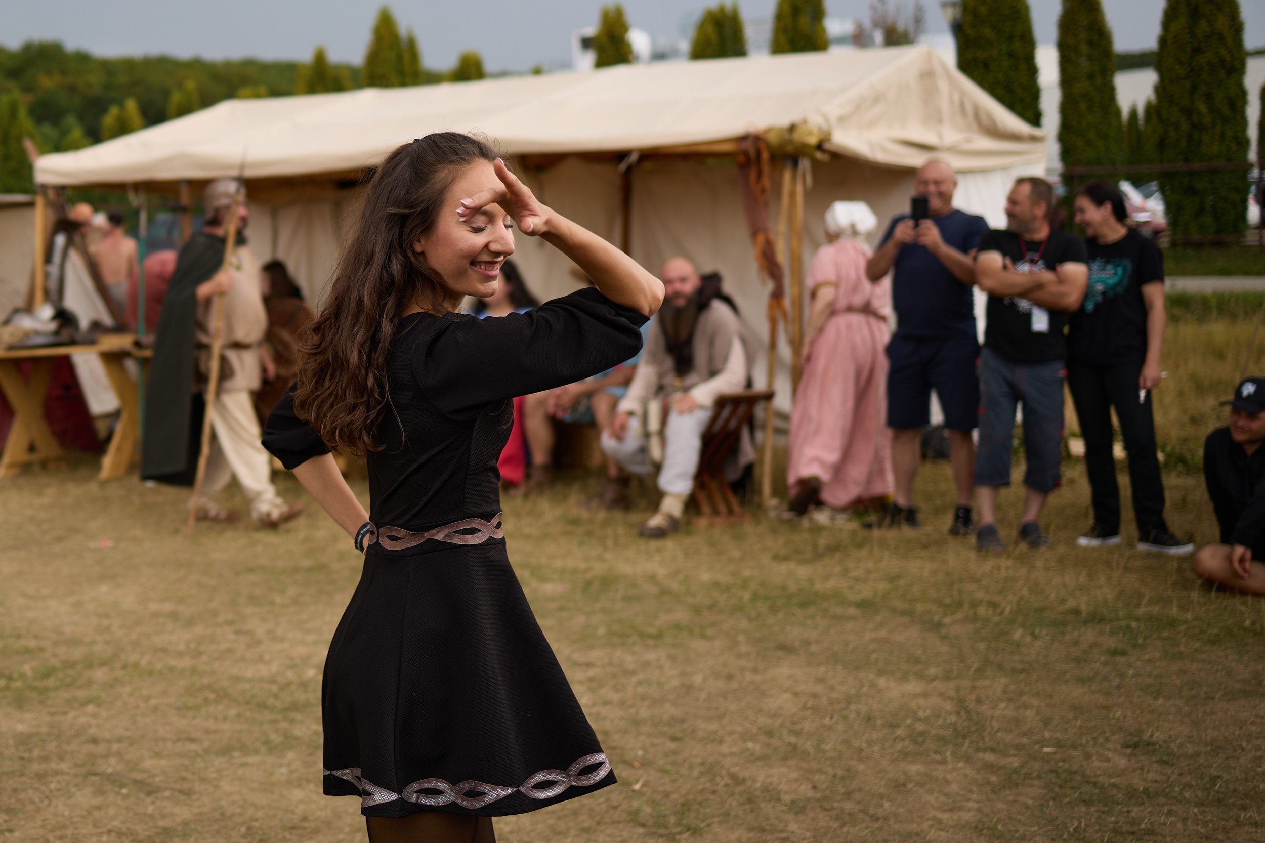 A smiling young woman with long brown hair, dressed in a black dress with silver accents, standing outdoors at a gathering with a beige canopy in the background. Several people are socializing and taking photos, and green trees are visible behind the