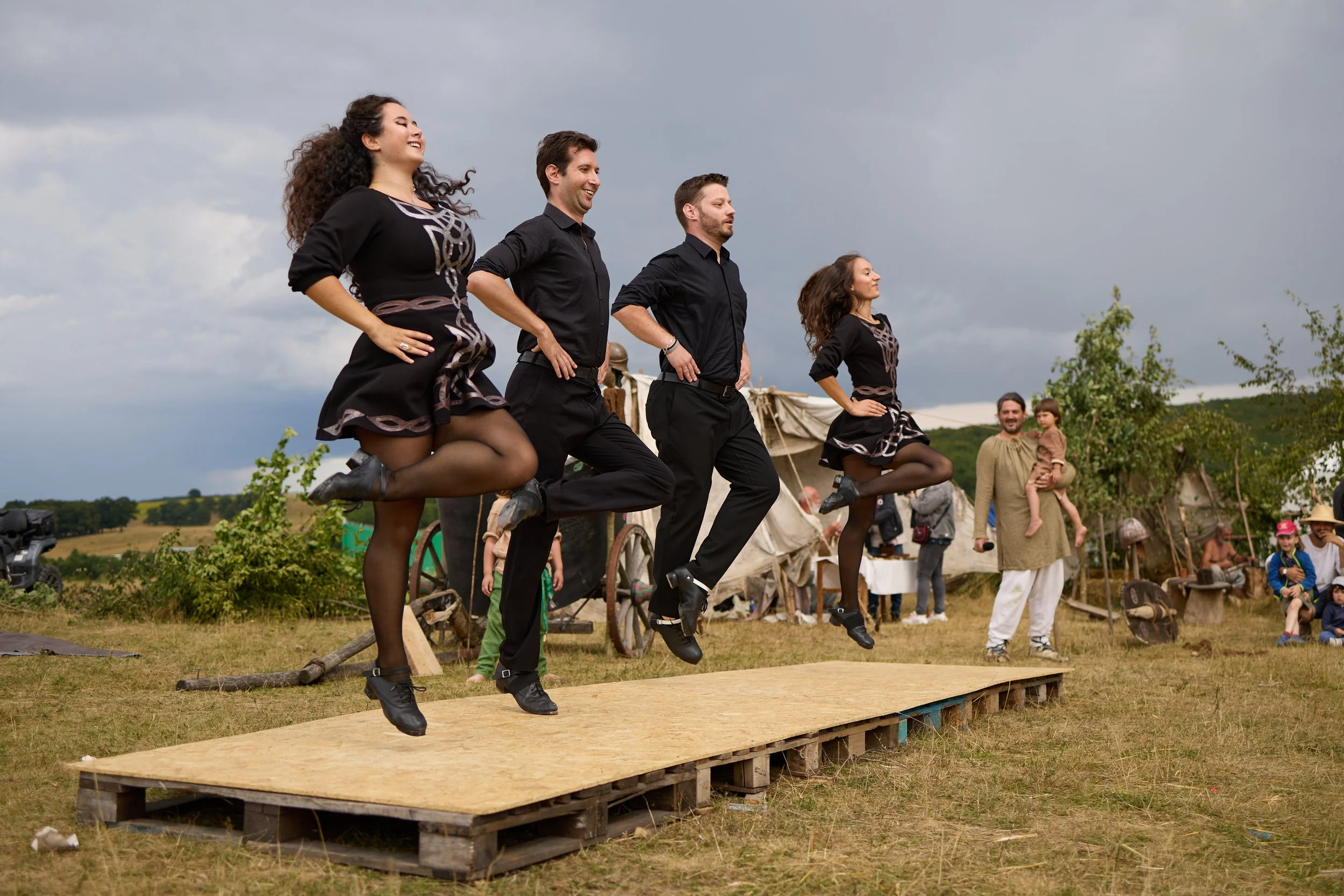 Four performers in black costumes with dance shoes perform a synchronized jump on a wooden platform outdoors during a cultural event, with a cloudy sky and audience in the background.
