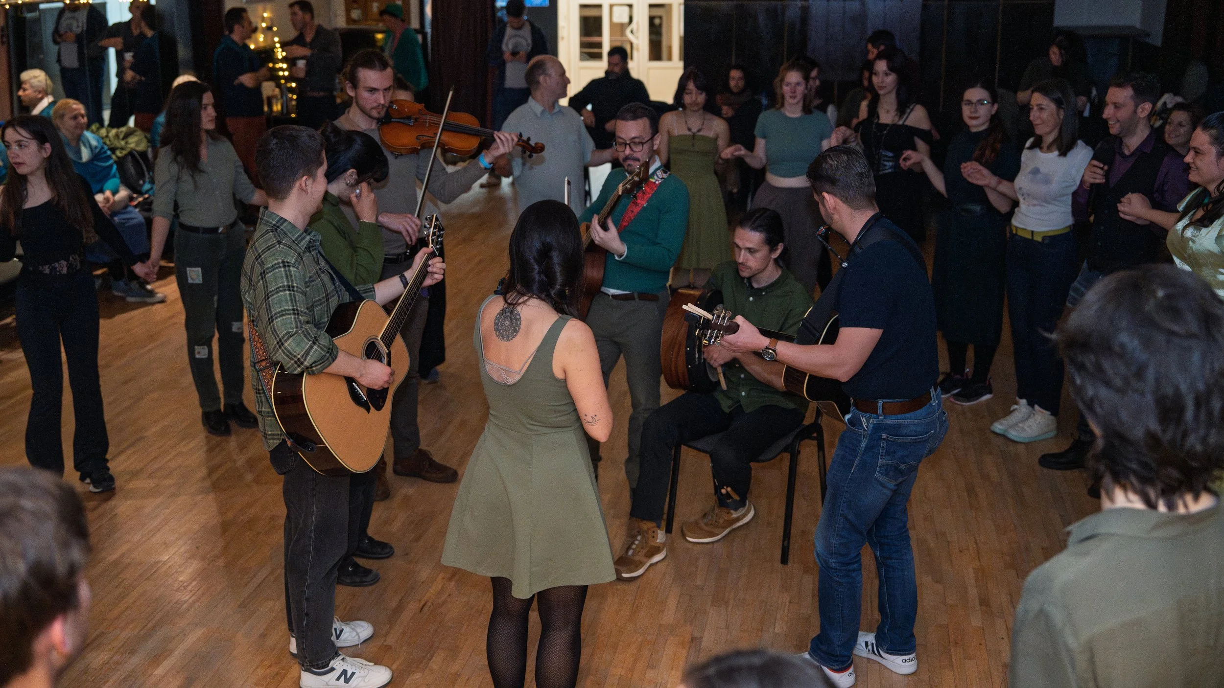 People gather in a room for a live music performance, some playing instruments like guitars, violin, and vocals, while others dance. The atmosphere is lively and social.