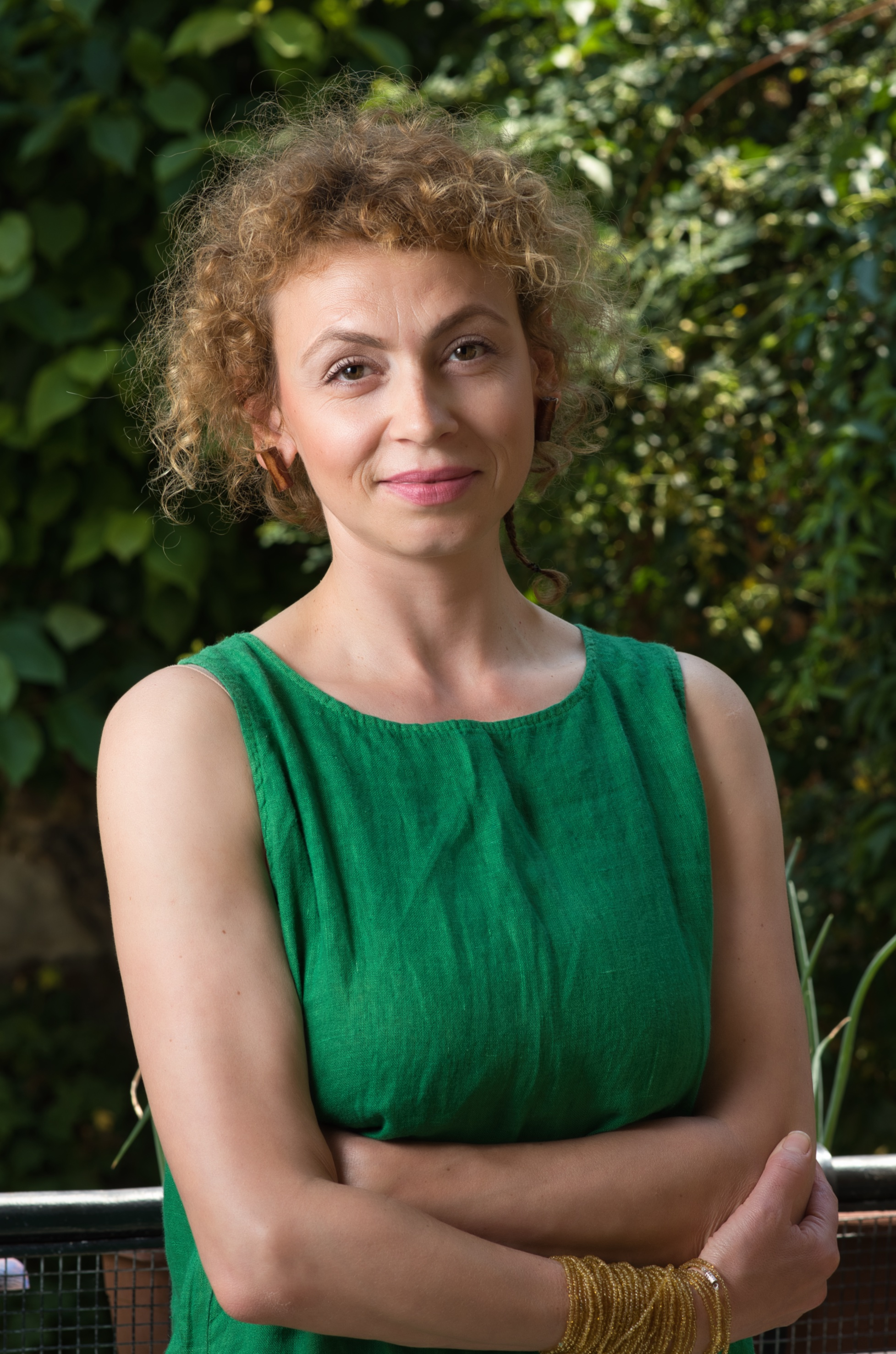 A woman with curly red hair wearing a green sleeveless top, standing outdoors with green foliage in the background.