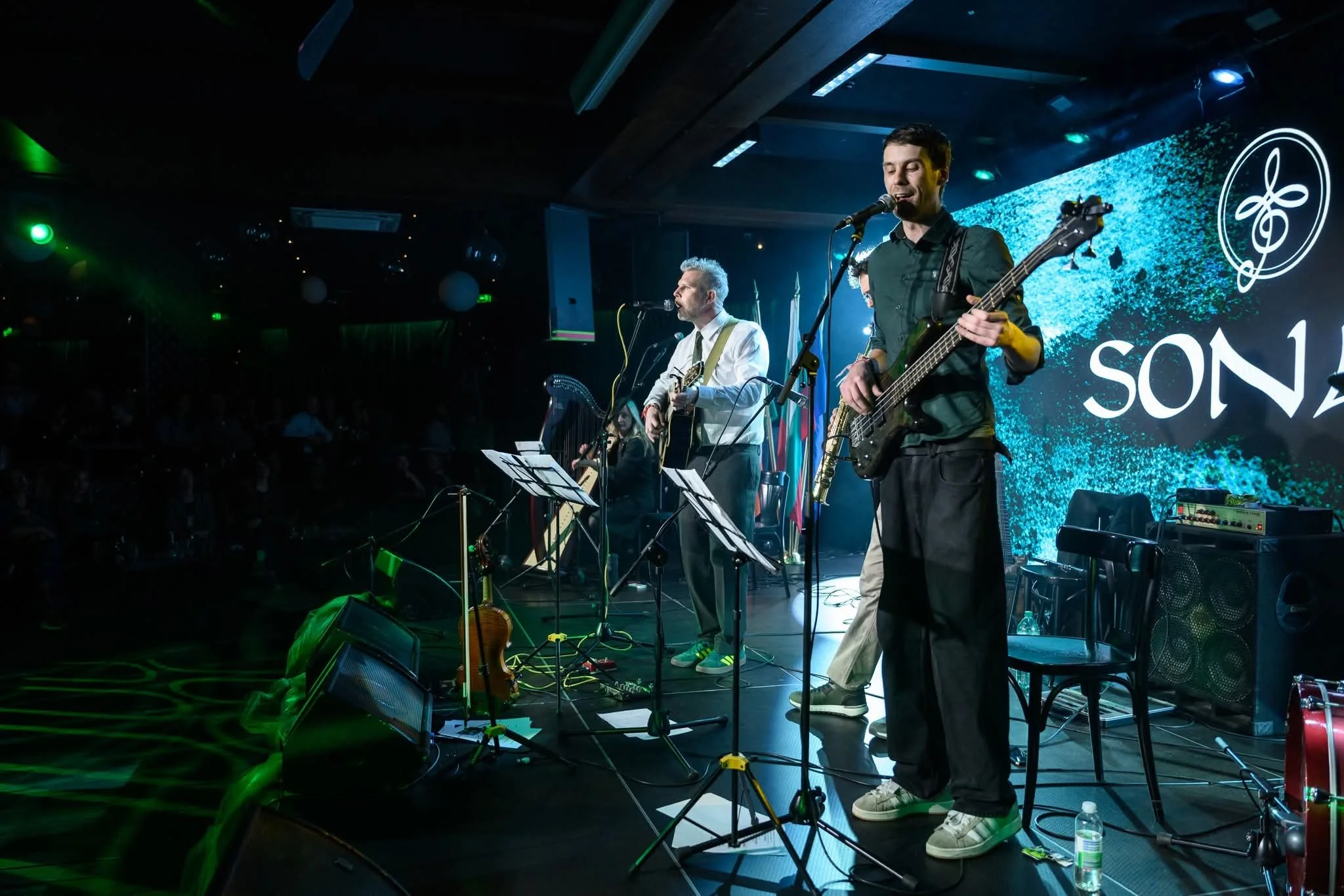 A band performs on stage with two musicians visible, one playing a guitar and singing, and the other playing a bass guitar. The stage is lit with colorful lighting, and a digital screen with a logo and the word 'SON' is in the background. The audienc