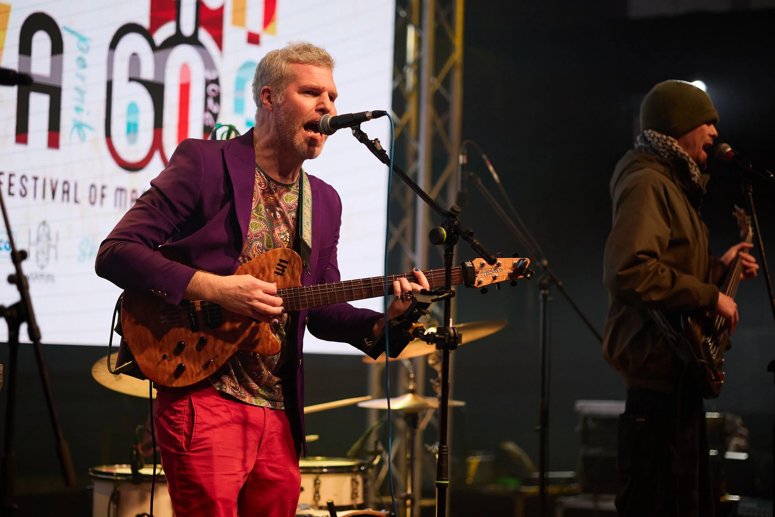 A musician with gray hair, beard, and wearing a purple blazer and red pants, singing into a microphone while playing an acoustic guitar during a live performance at a festival, with a large screen and another musician in winter clothing in the backgr